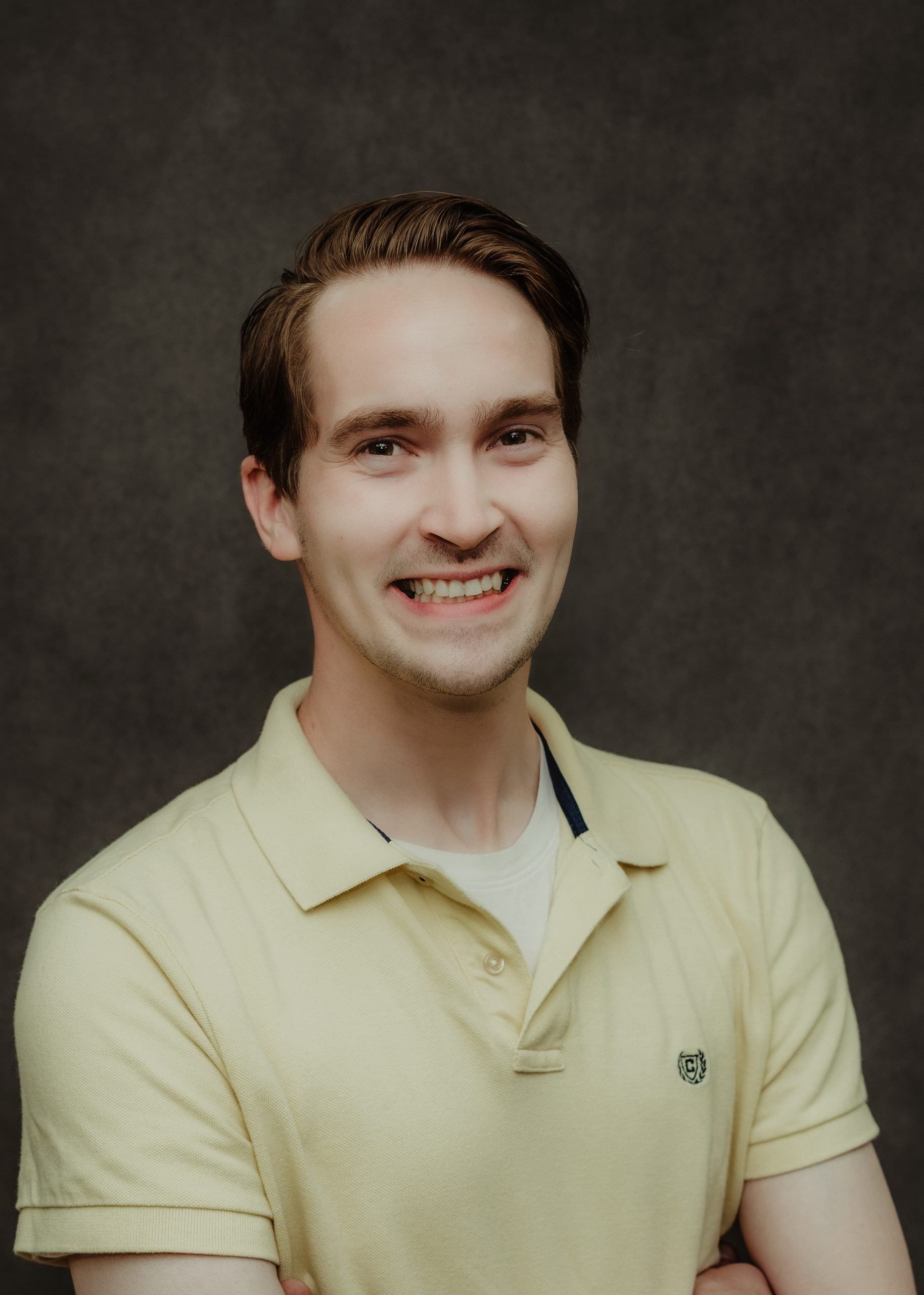A person with brown hair smiles, wearing a light yellow polo shirt against a dark gray background.