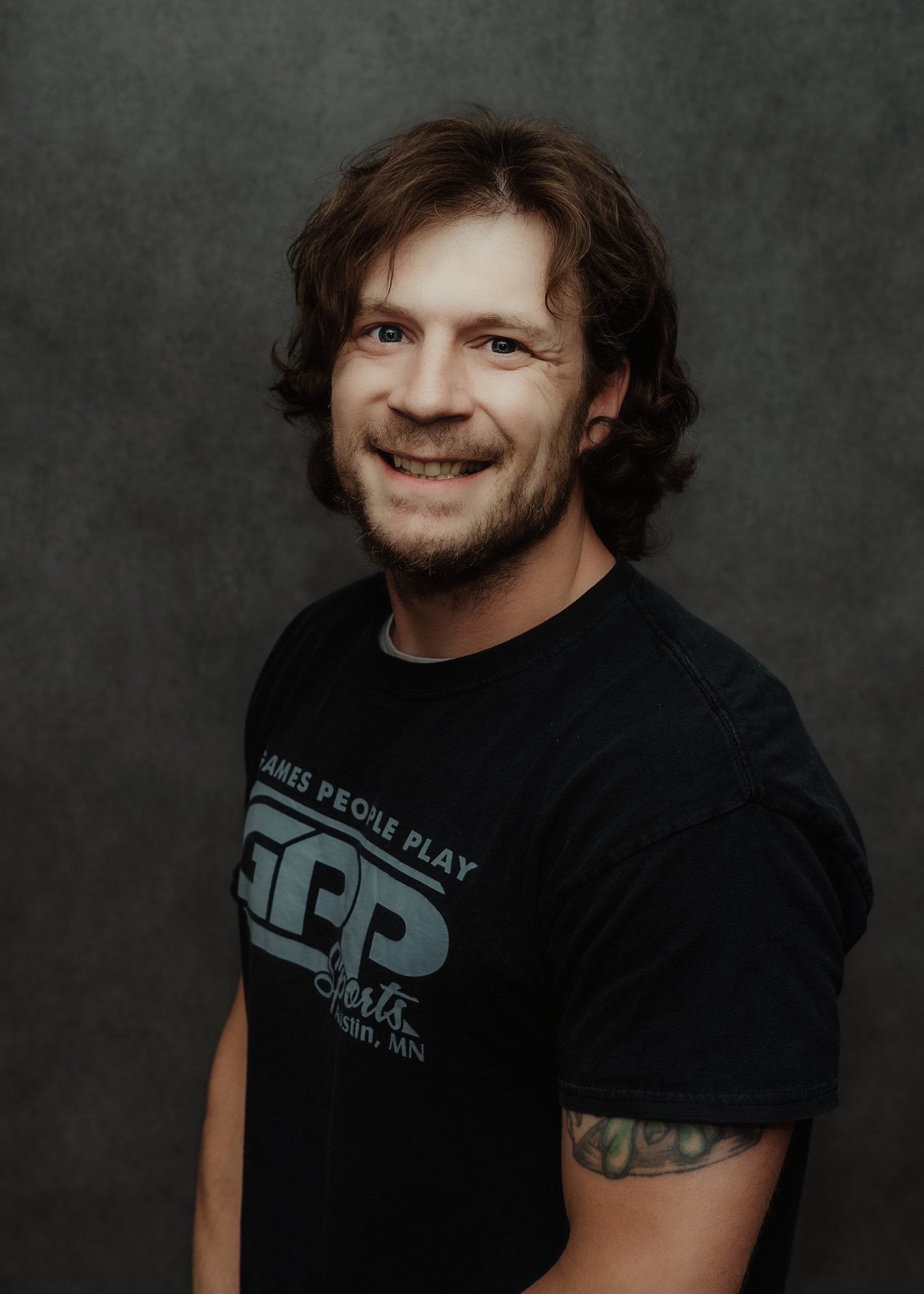 A smiling person with wavy brown hair wearing a dark t-shirt with a logo, set against a dark grey background.