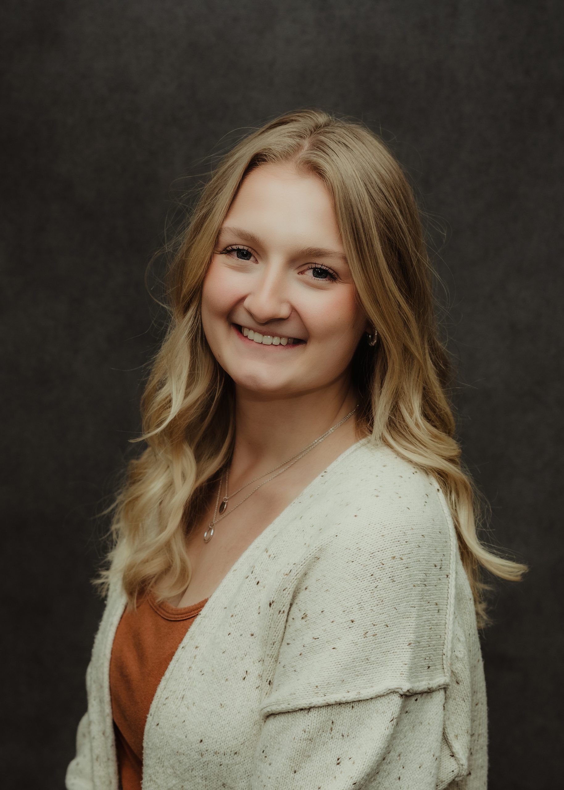 A smiling person with long blonde hair, wearing a white textured cardigan over a rust-colored top against a dark background.