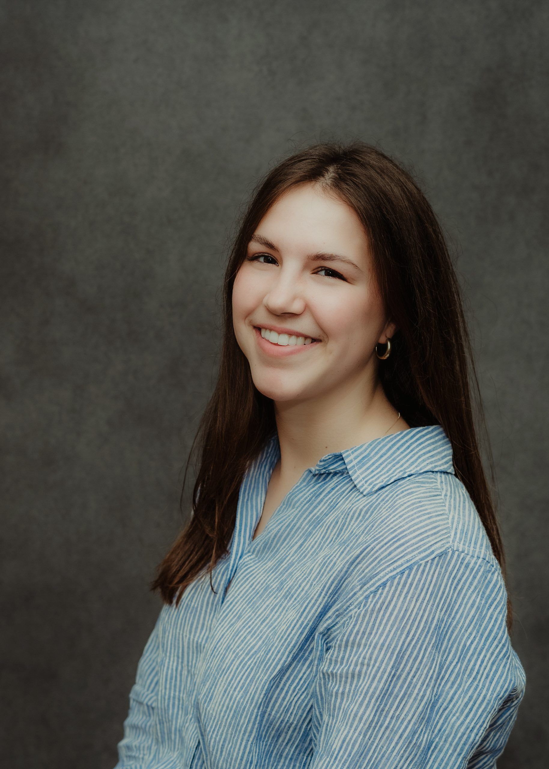 A person with long, brown hair, wearing a light blue, collared striped shirt, smiles against a dark, mottled background.