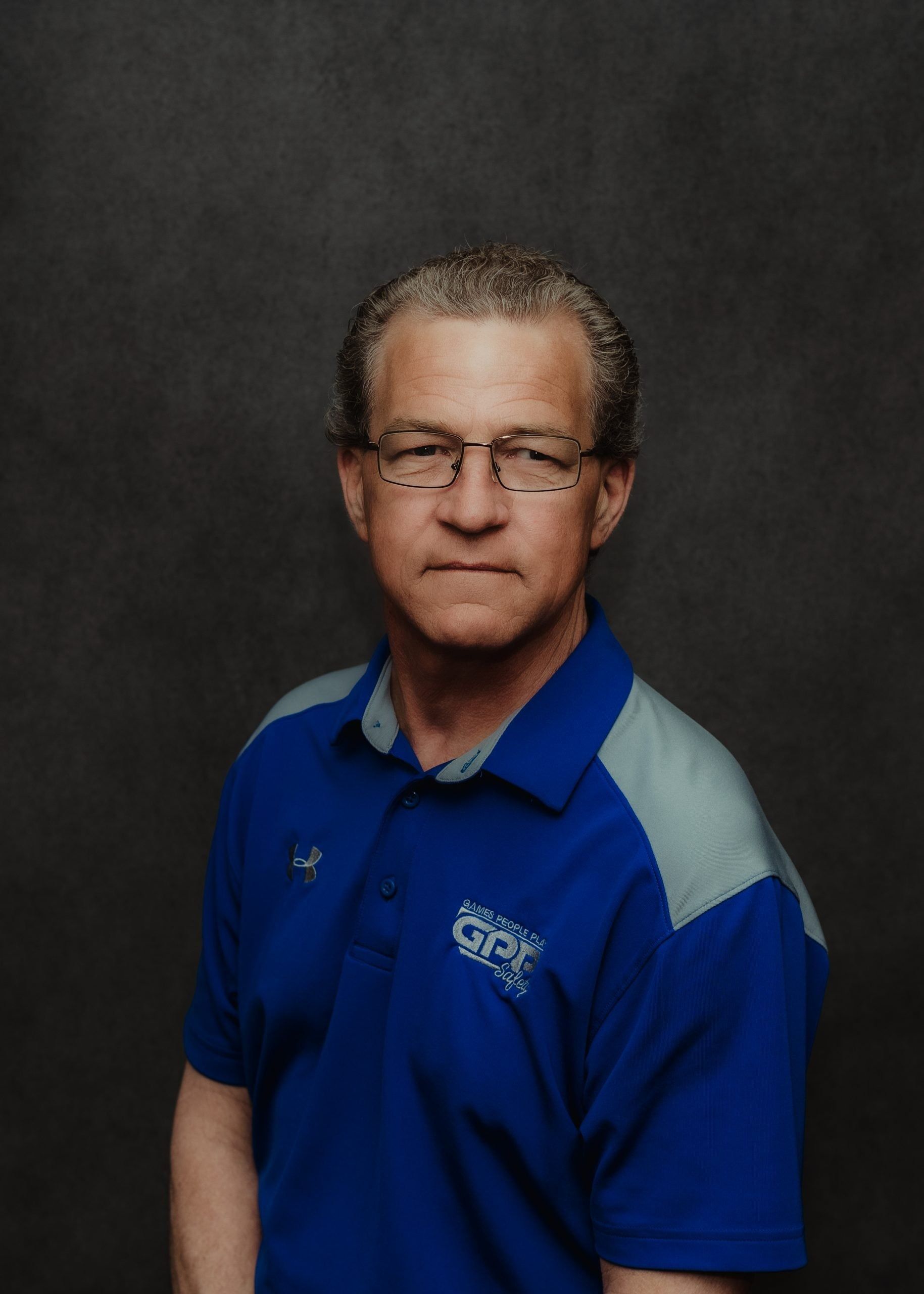 A person wearing a blue polo shirt with grey shoulders and a logo, standing against a dark grey background.