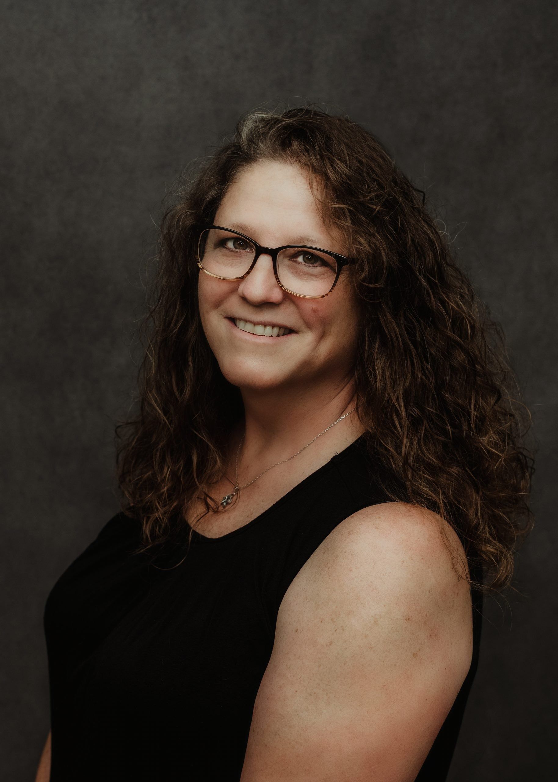 A person with long, curly hair and glasses smiles in a headshot against a dark, textured gray background.