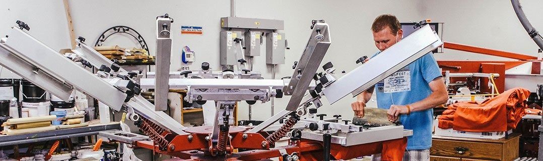 A person wearing a blue t-shirt operates a large, multi-arm screen printing press in a workshop.
