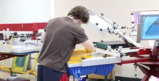 A person in a brown t-shirt operates a screen printing press, manually applying ink onto a blue fabric garment.