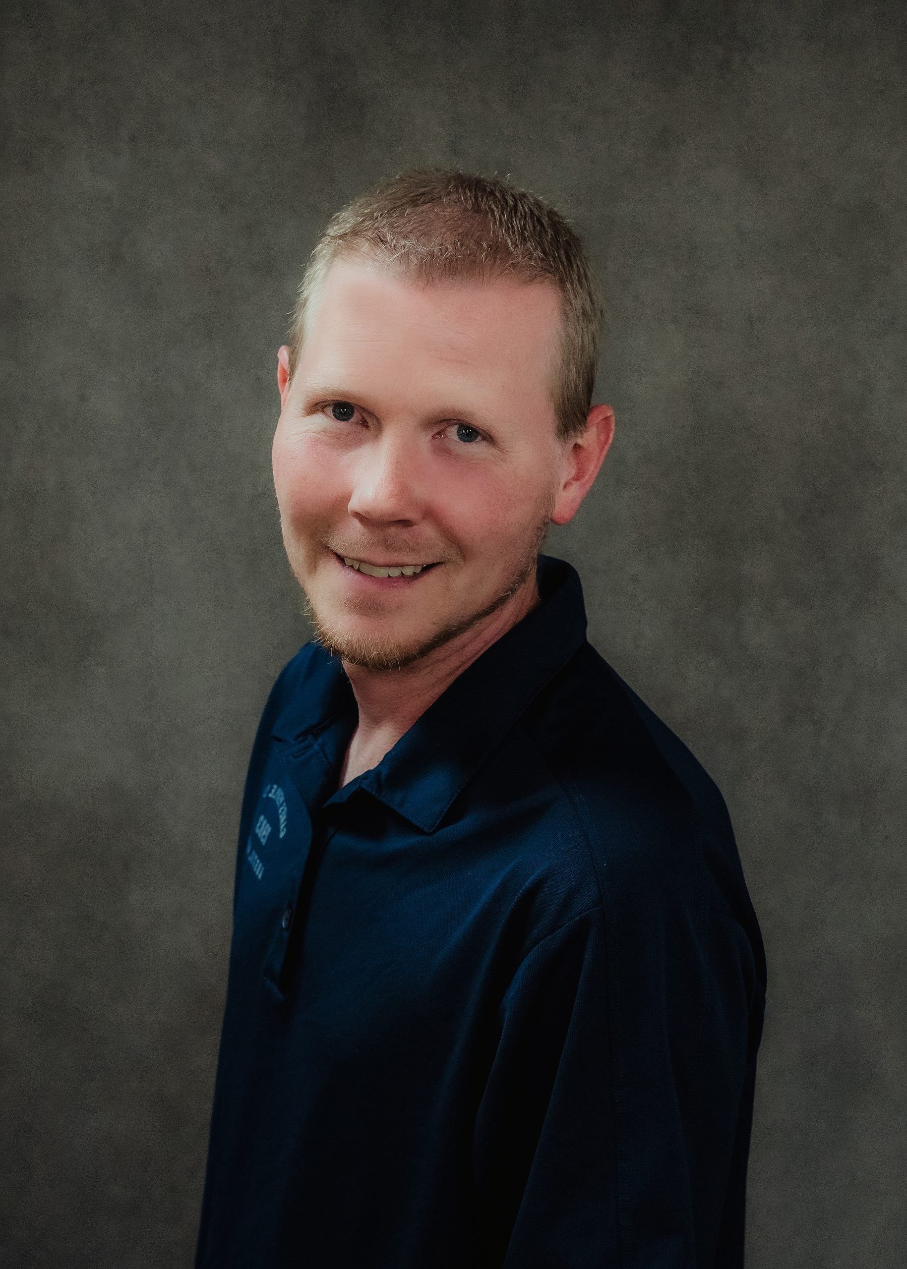 A person wearing a dark blue polo shirt against a textured gray background, smiling slightly.