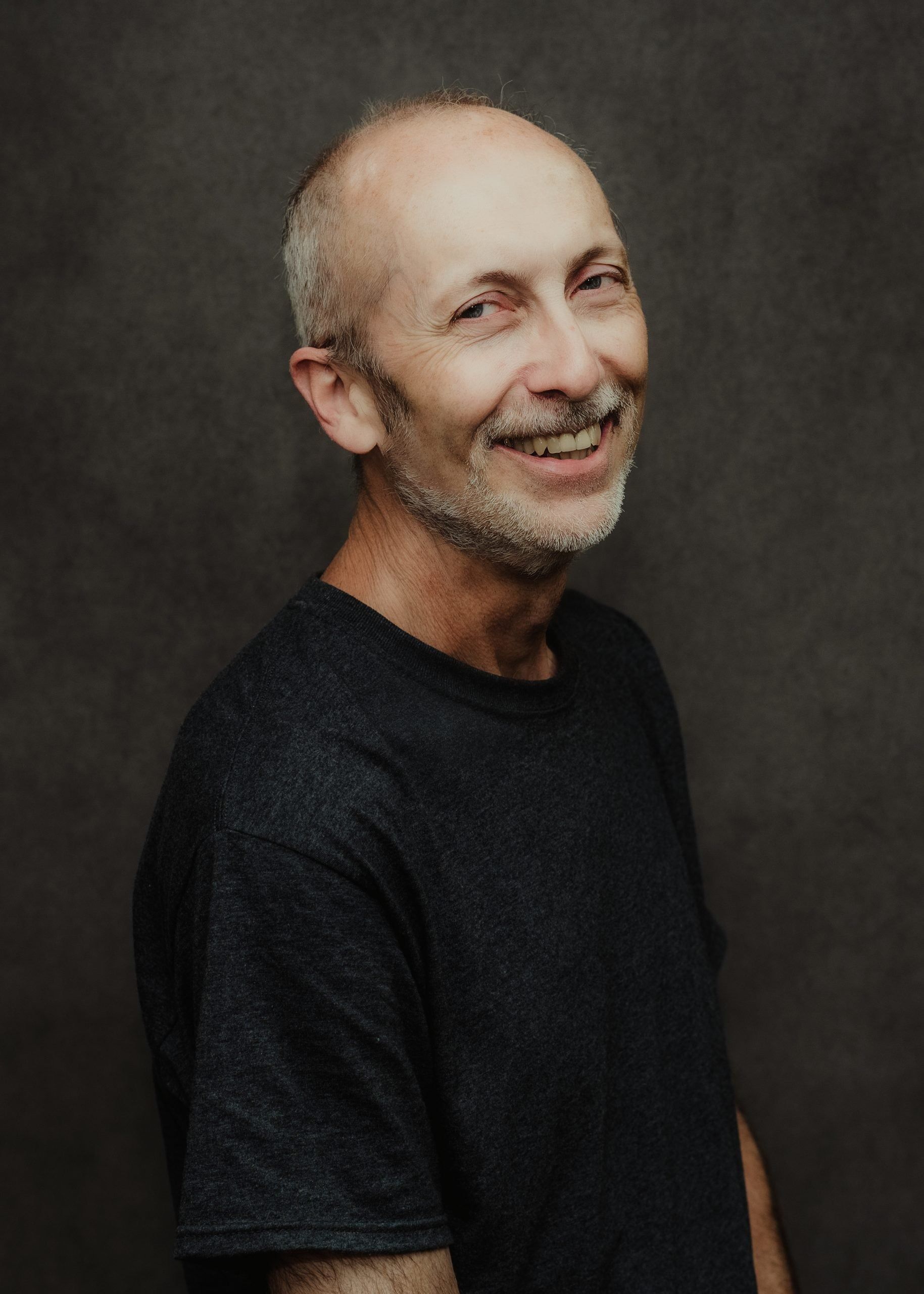 A man with a short beard smiles while looking at the camera against a dark, textured gray background.