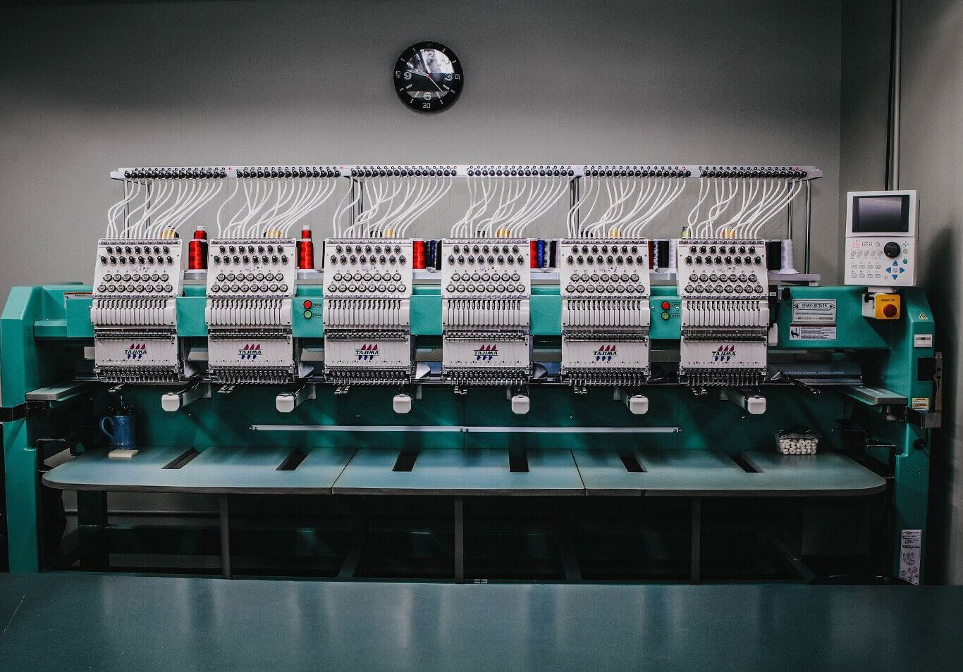 A large, teal, multi-head industrial embroidery machine with many spools of thread sits in a workshop.