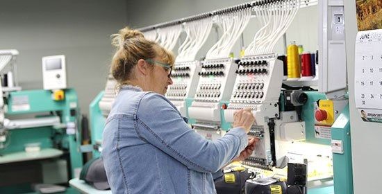 A person operating a multi-head embroidery machine in a workshop, adjusting settings on the control panel.