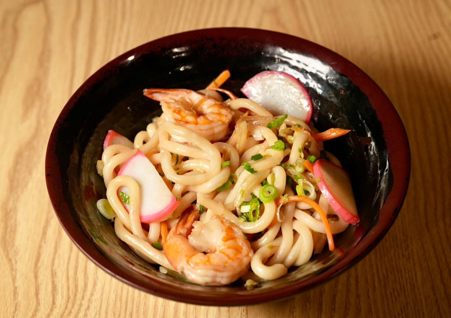 udon noodles with shrimp, fish cakes, and vegetables in a dark bowl on a wooden surface