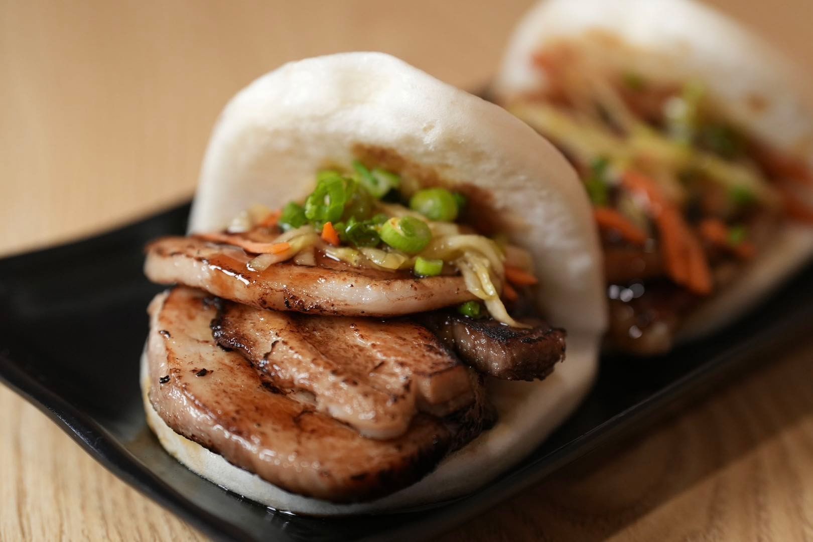 two steamed bao buns filled with grilled pork belly, and pickled vegetables on a black plate