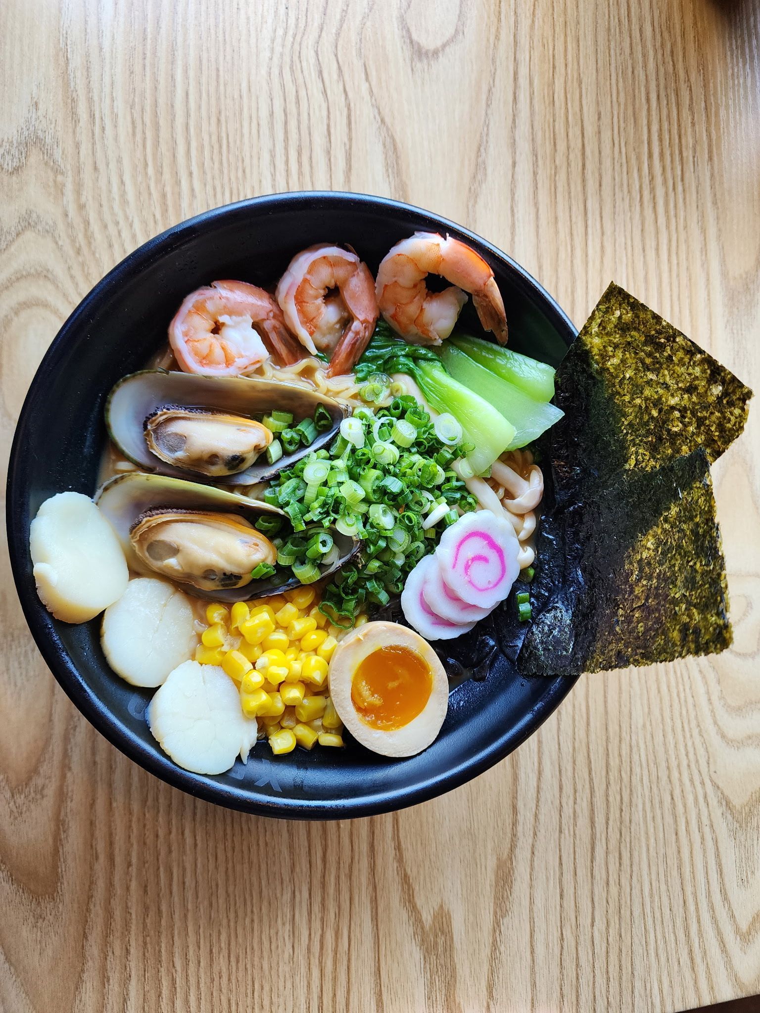 ramen bowl with shrimp, mussels, egg, corn, vegetables, and seaweed on a wooden table