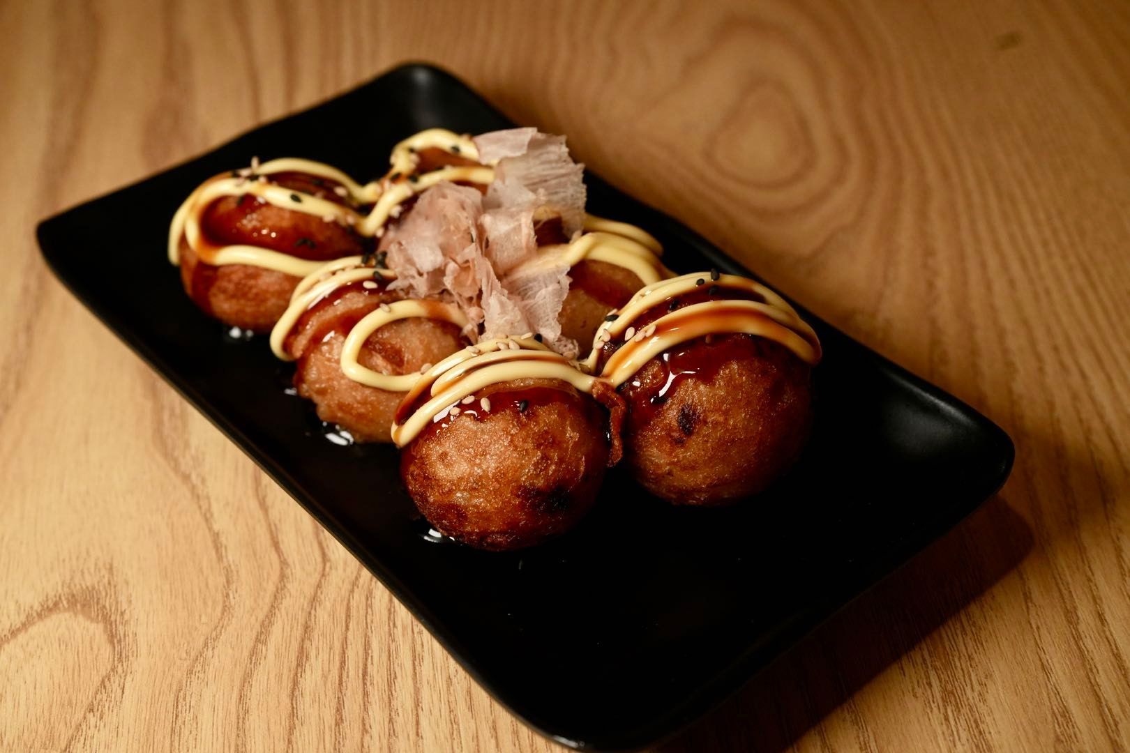 takoyaki balls on a black plate, drizzled with sauce and mayo, topped with bonito flakes, on a wooden table