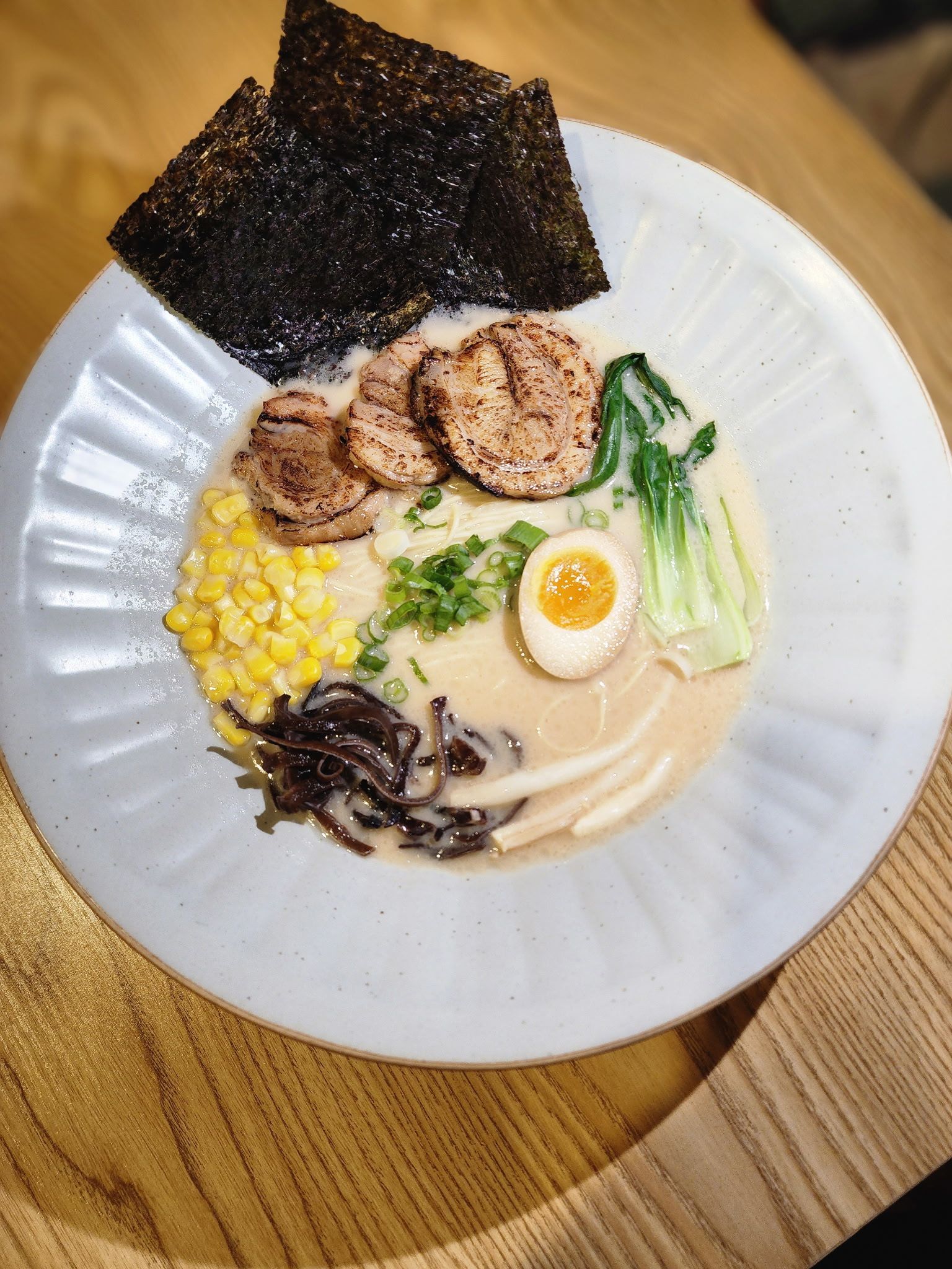 ramen bowl with pork, egg, seaweed, corn, and bok choy in a creamy broth on a wooden table