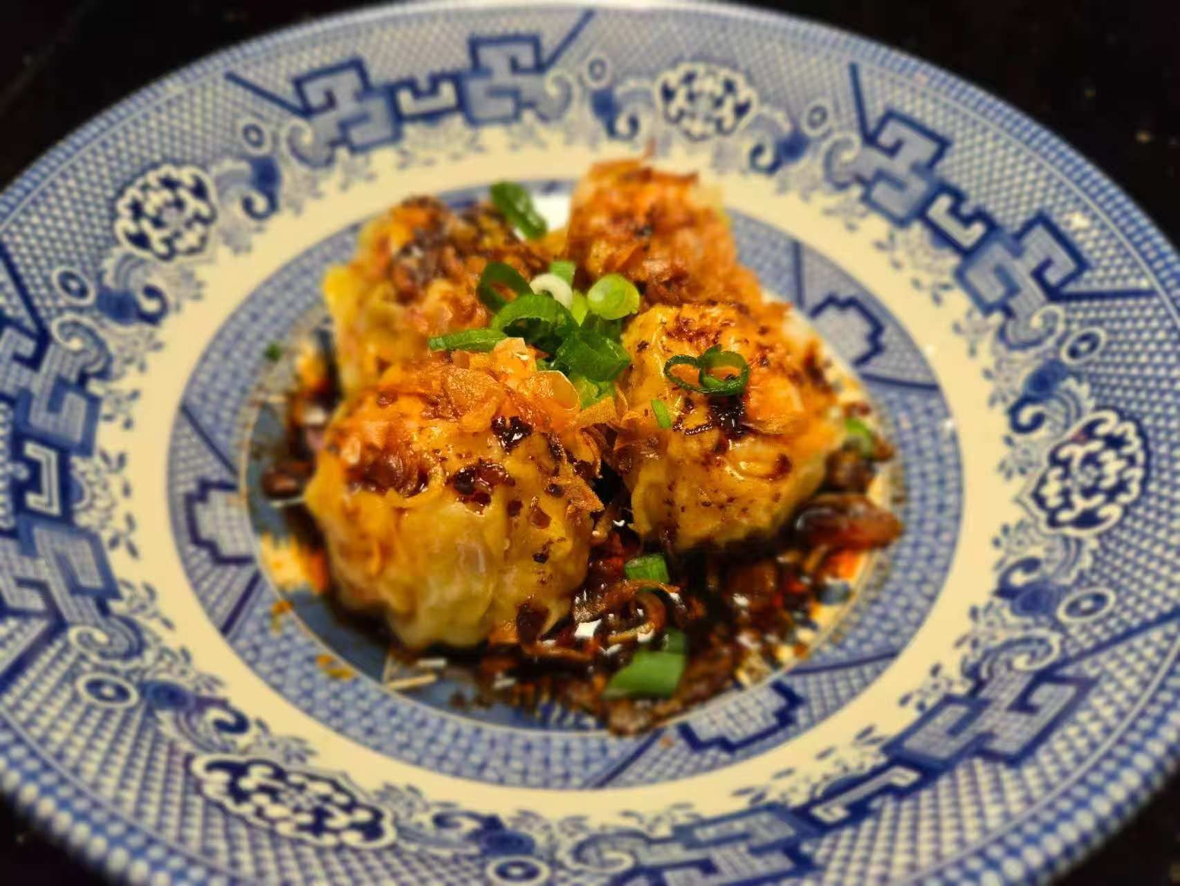 steamed dumplings in a spicy sauce, garnished with green onions, on a blue and white patterned plate