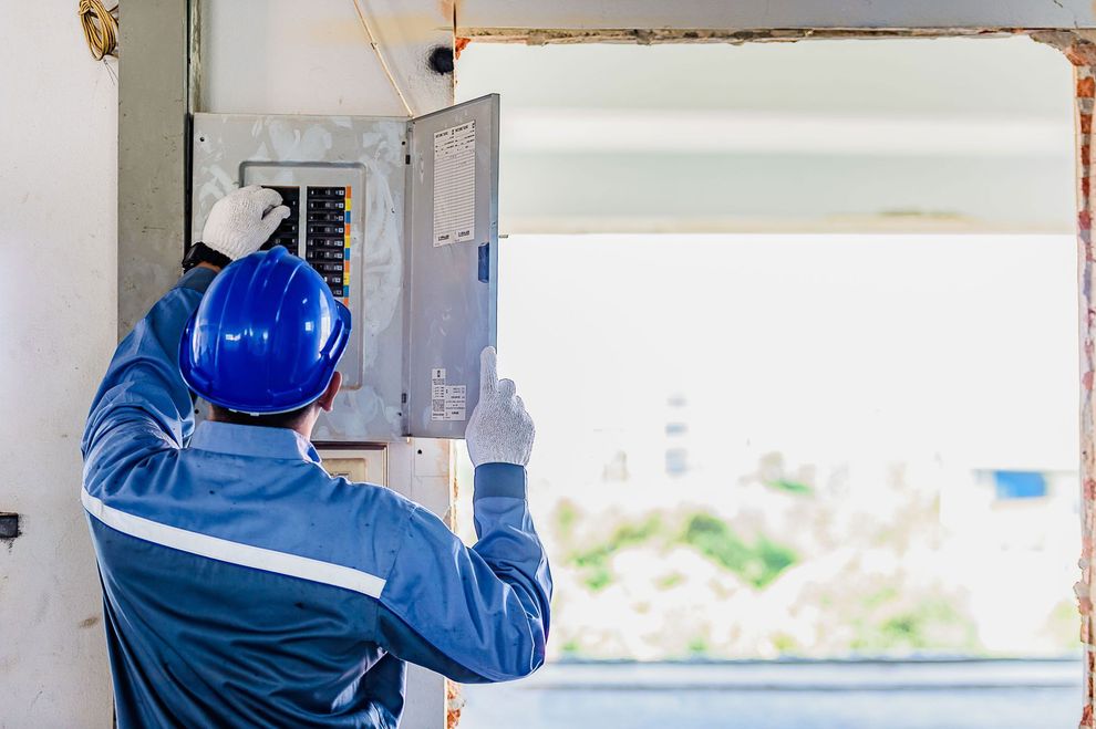 Electrician in blue uniform and hard hat working on a circuit breaker panel.