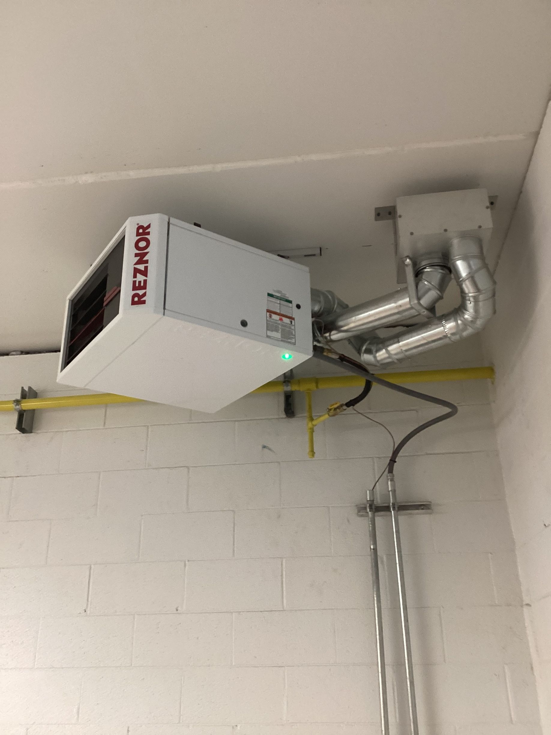 A Reznor ceiling-mounted gas heater with attached metal exhaust pipes, installed against a white brick wall.
