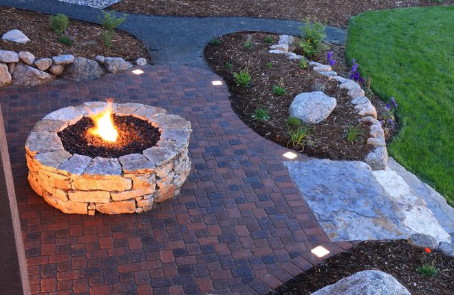 Fire pit in a brick patio, surrounded by landscaped gardens and path, illuminated at dusk.