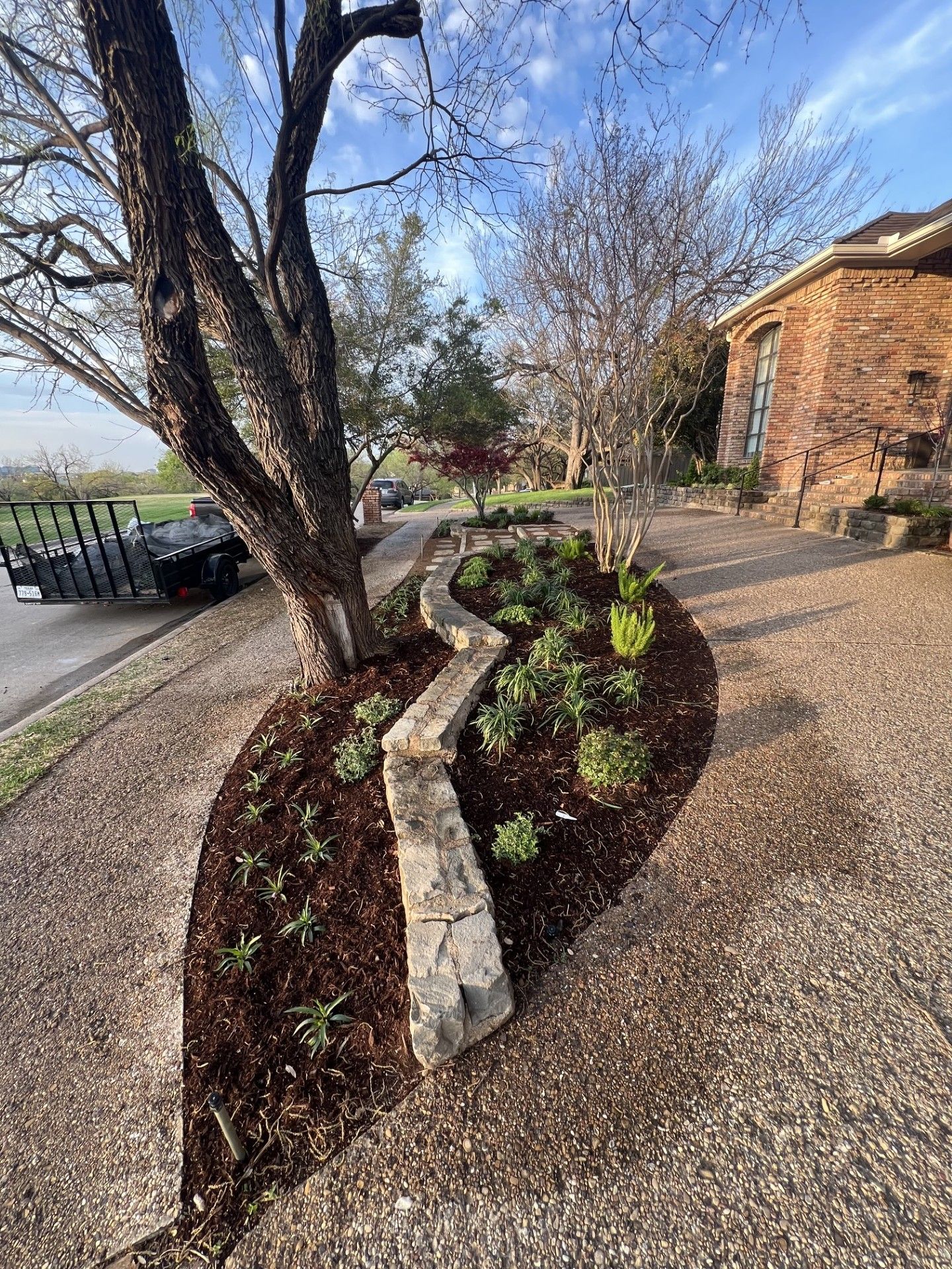Landscaped garden bed with stone edging beside a house and driveway, bordered by trees and shrubs