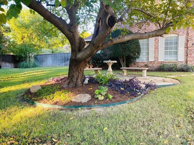 Backyard with a large tree, flower bed, birdbath, and a brick house in the background
