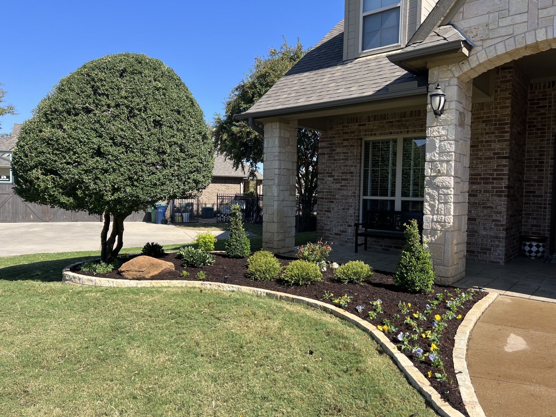 Front yard with trimmed tree, brick house porch, and curved flower bed under a clear blue sky