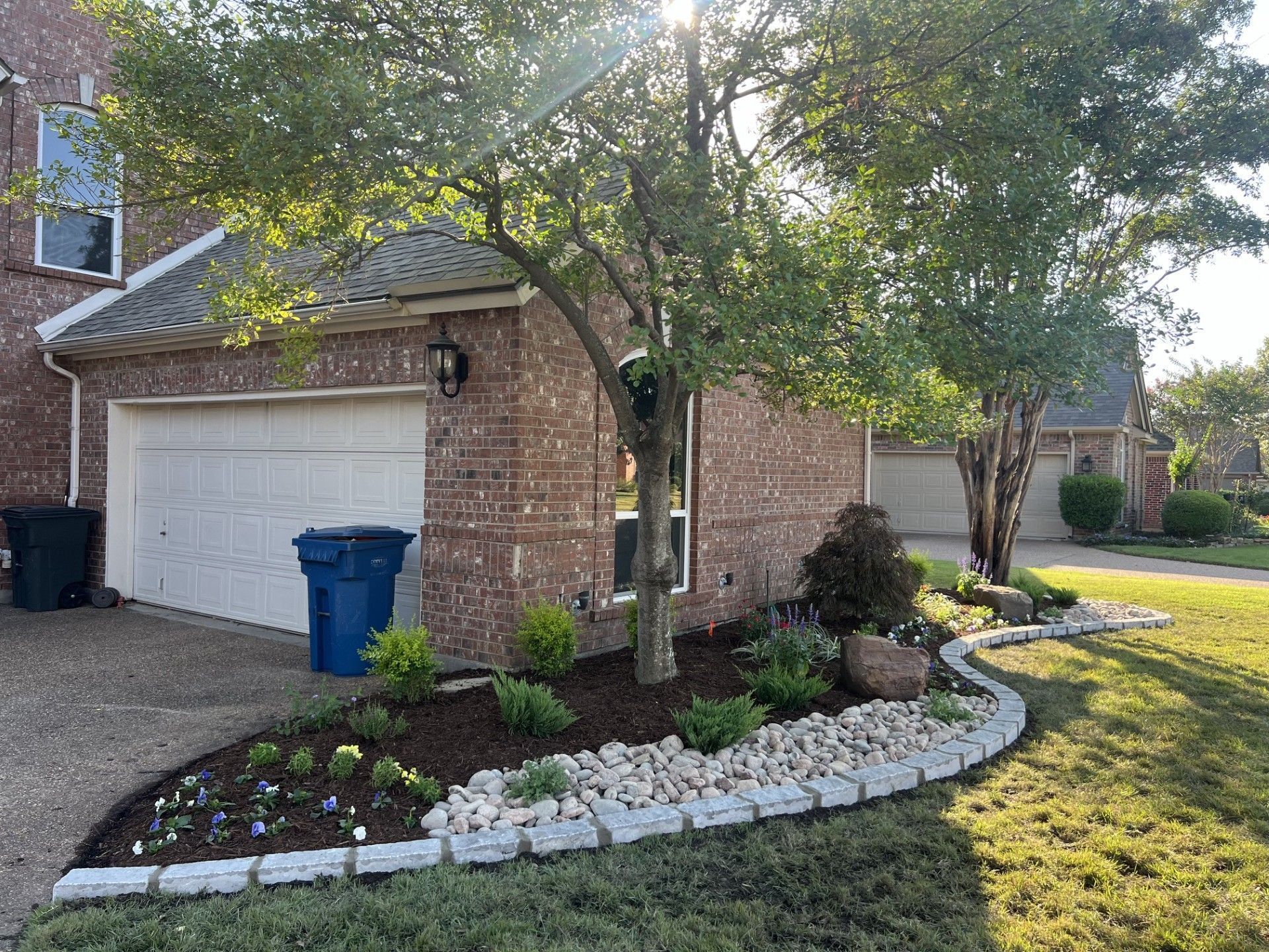 Suburban brick house with tree-shaded front yard, landscaped flower bed, and two blue trash bins by the garage.