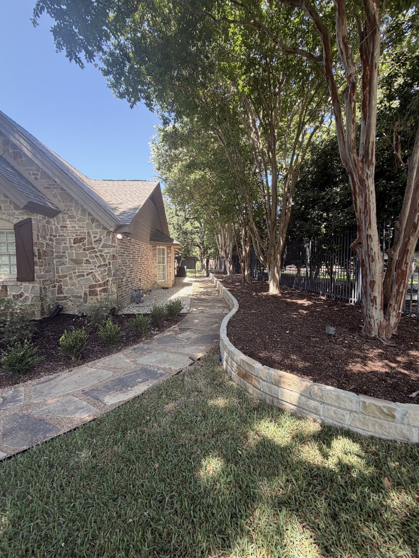 Stone house beside curving white wall and tree-lined walkway in a sunny garden courtyard