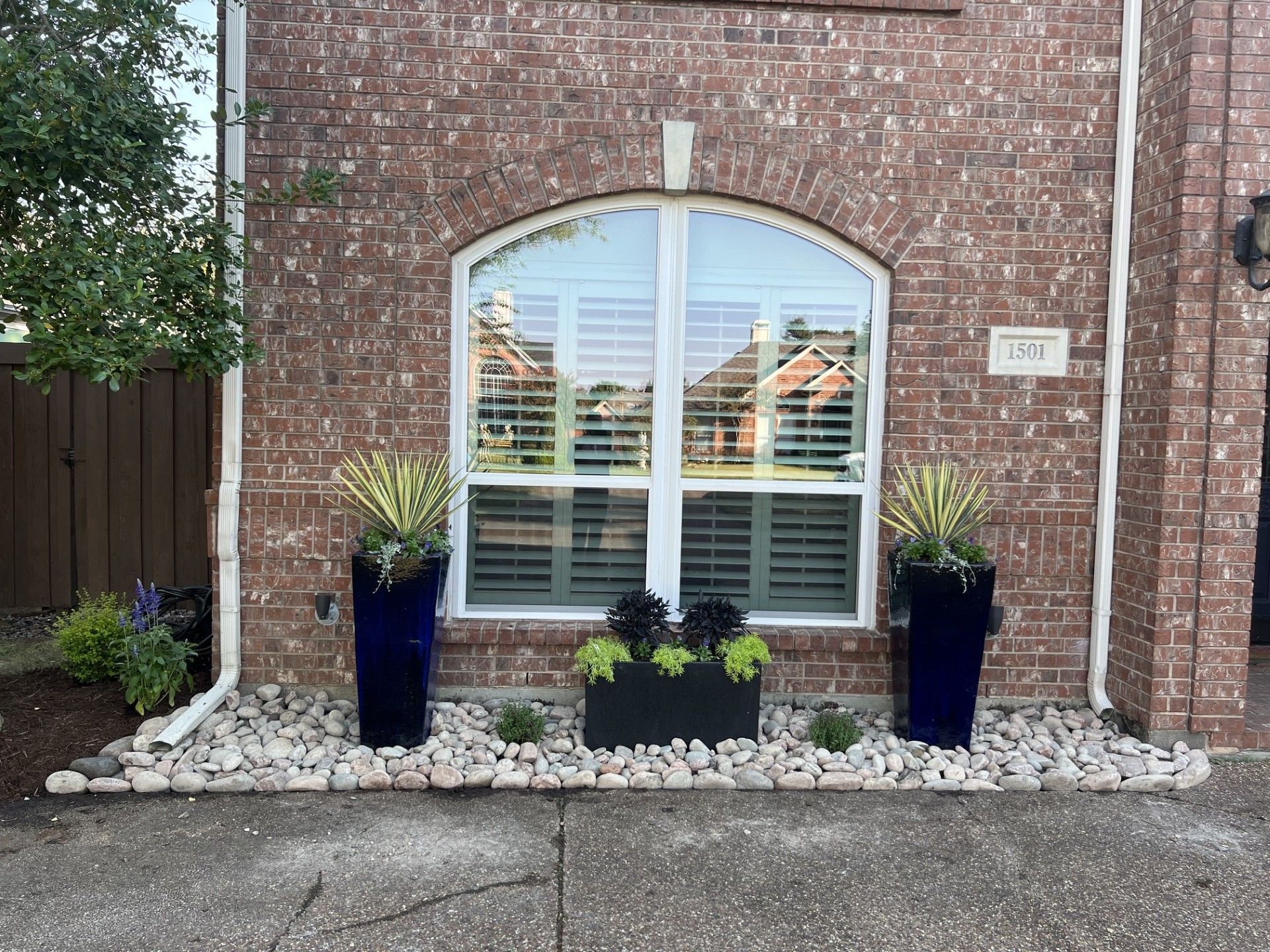 Brick house entry with arched window, white shutters, and potted plants on a gravel bed