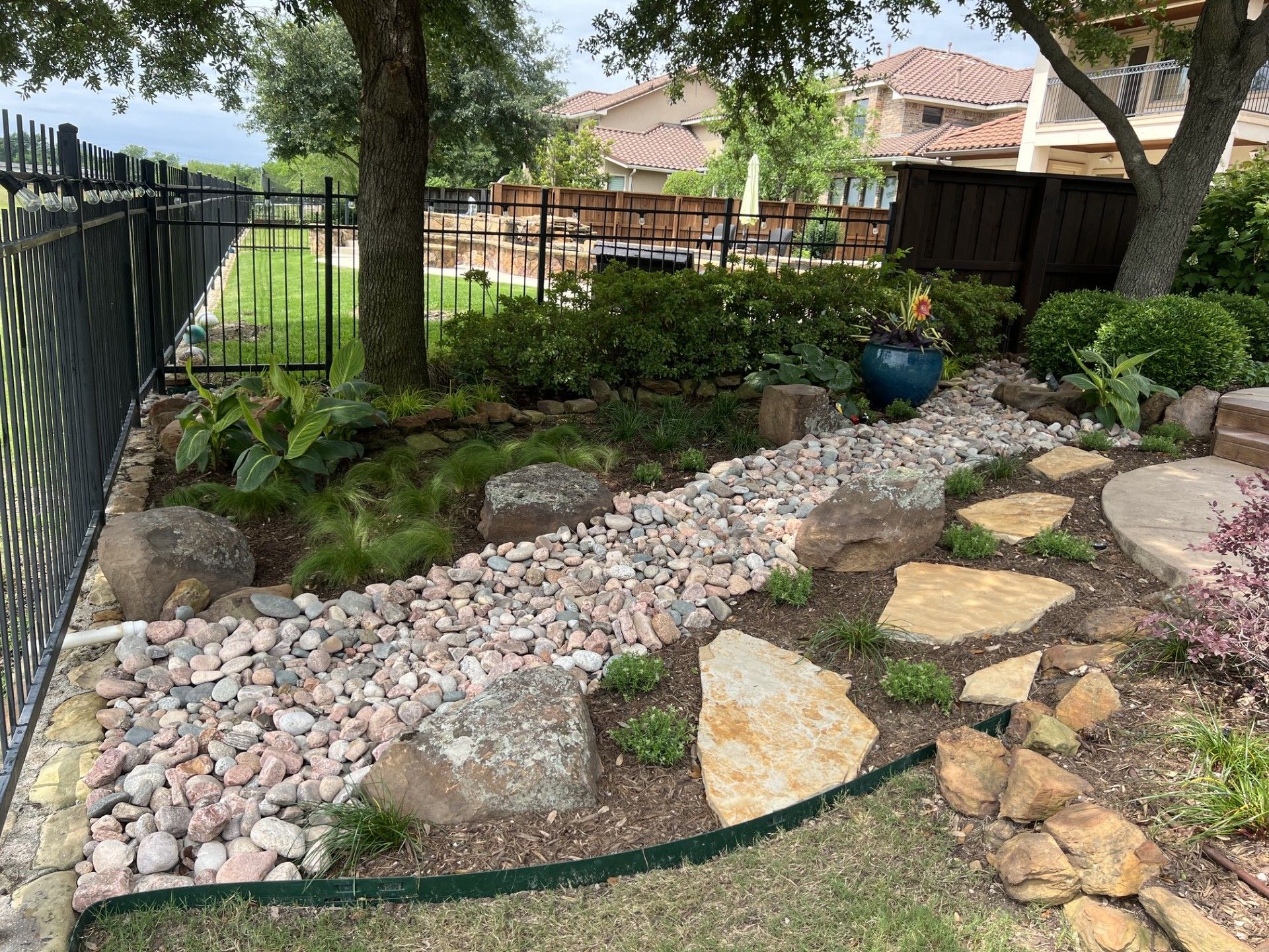 Backyard garden with rock-lined dry creek bed, stone path, trees, and a black fence