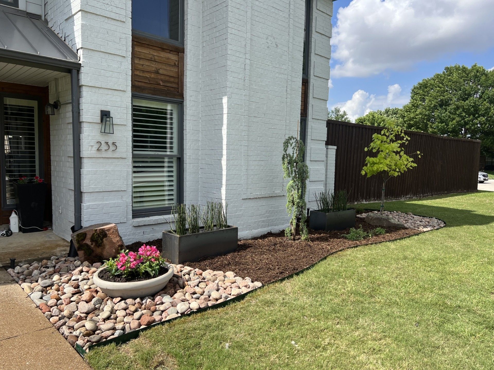 Front yard with white house, rock border, potted flowers, and green lawn beside a wooden fence.