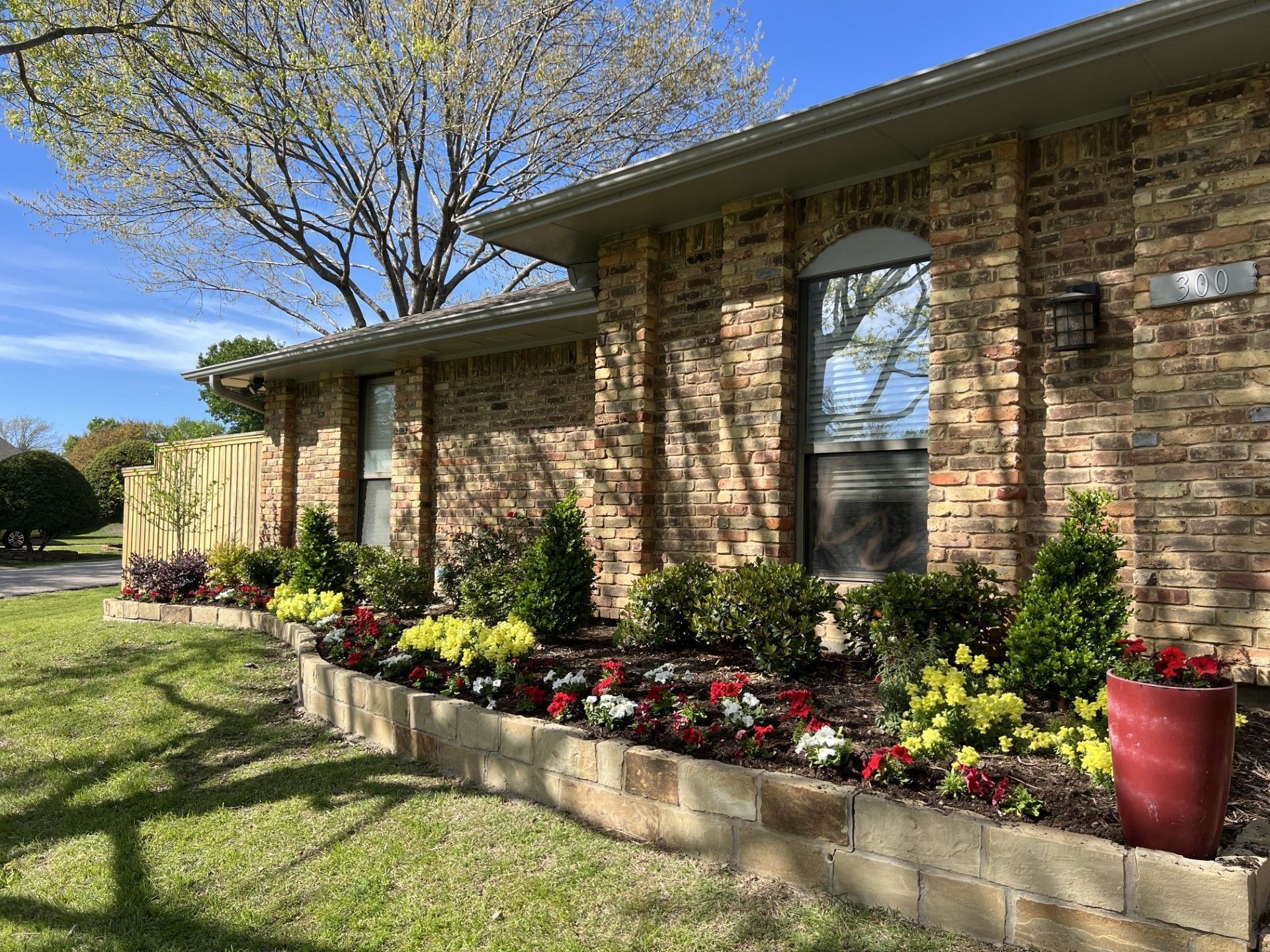 Brick house front with flower bed, potted plants, and a leafy tree in the yard
