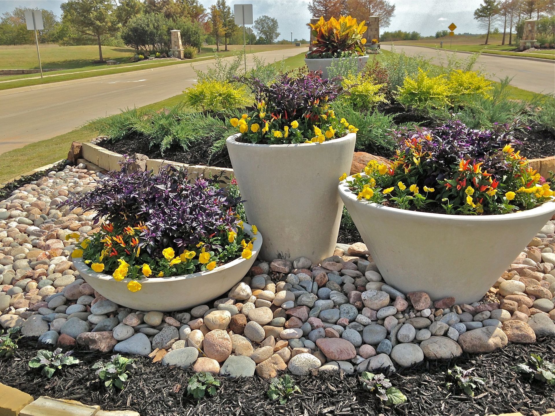 Three large planters with colorful flowers on a rock bed beside a roadside garden.