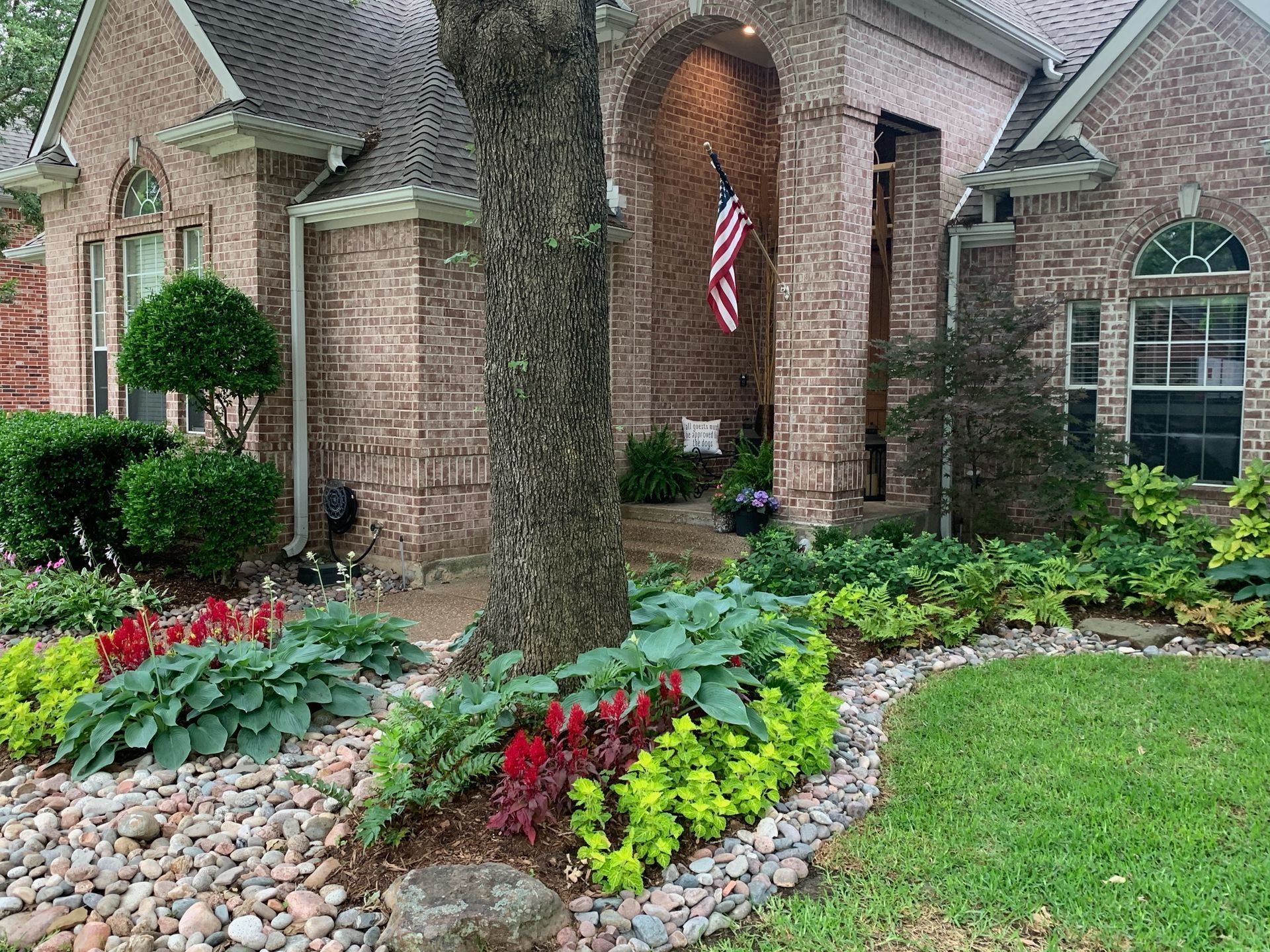 Front yard with brick house, curved stone border, colorful flowers, shrubs, and an American flag by the porch.