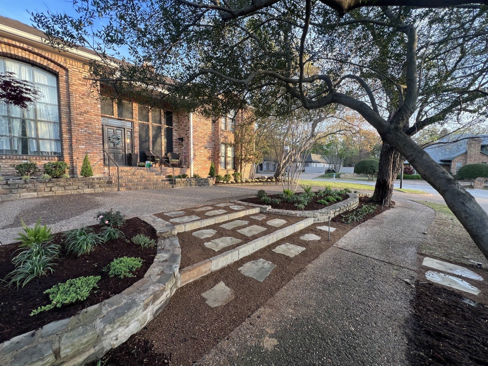 Brick courtyard walkway with stepping stones, garden beds, and a large tree beside a building