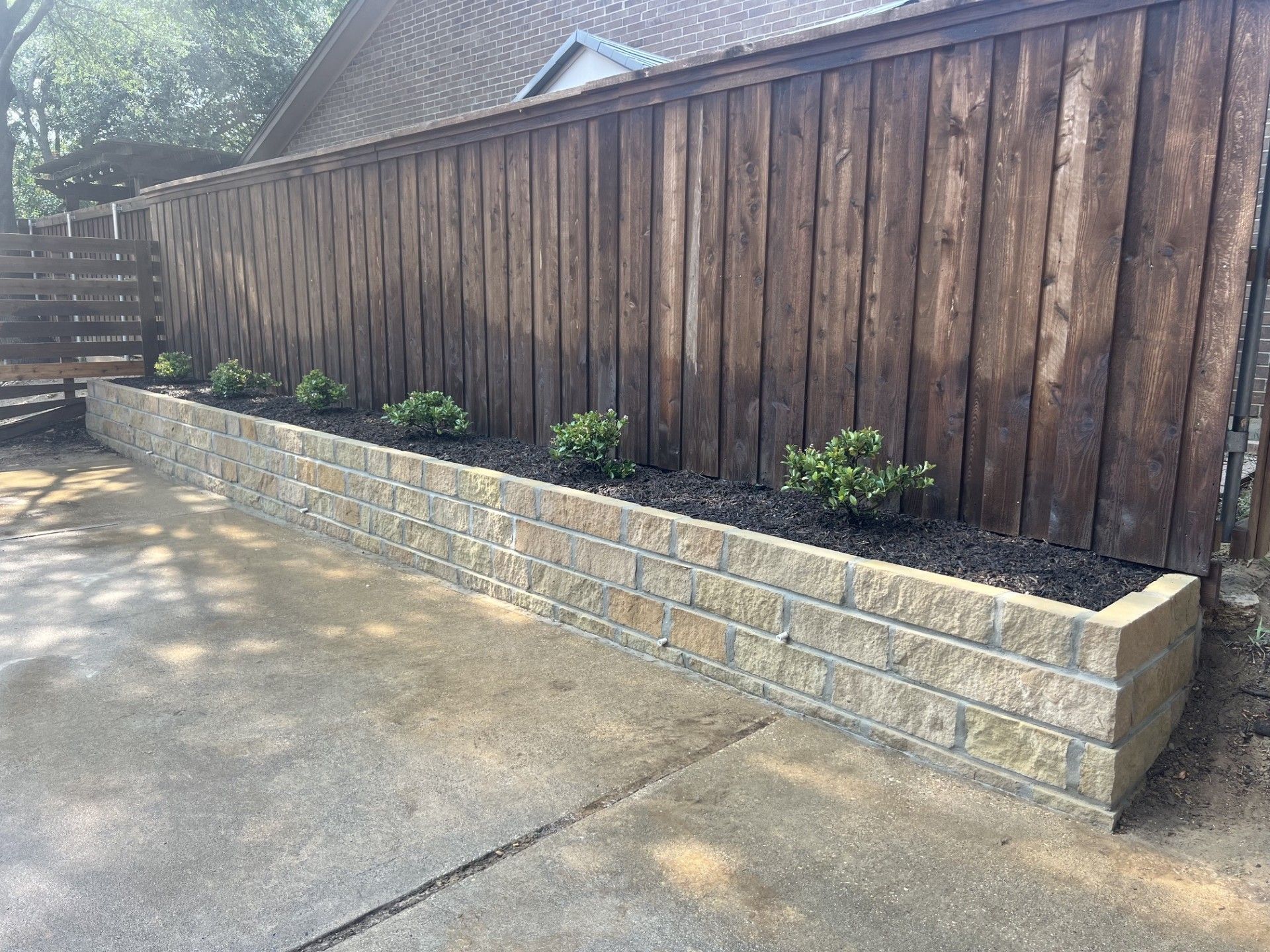 Wooden fence beside raised stone planter with small shrubs along a concrete driveway