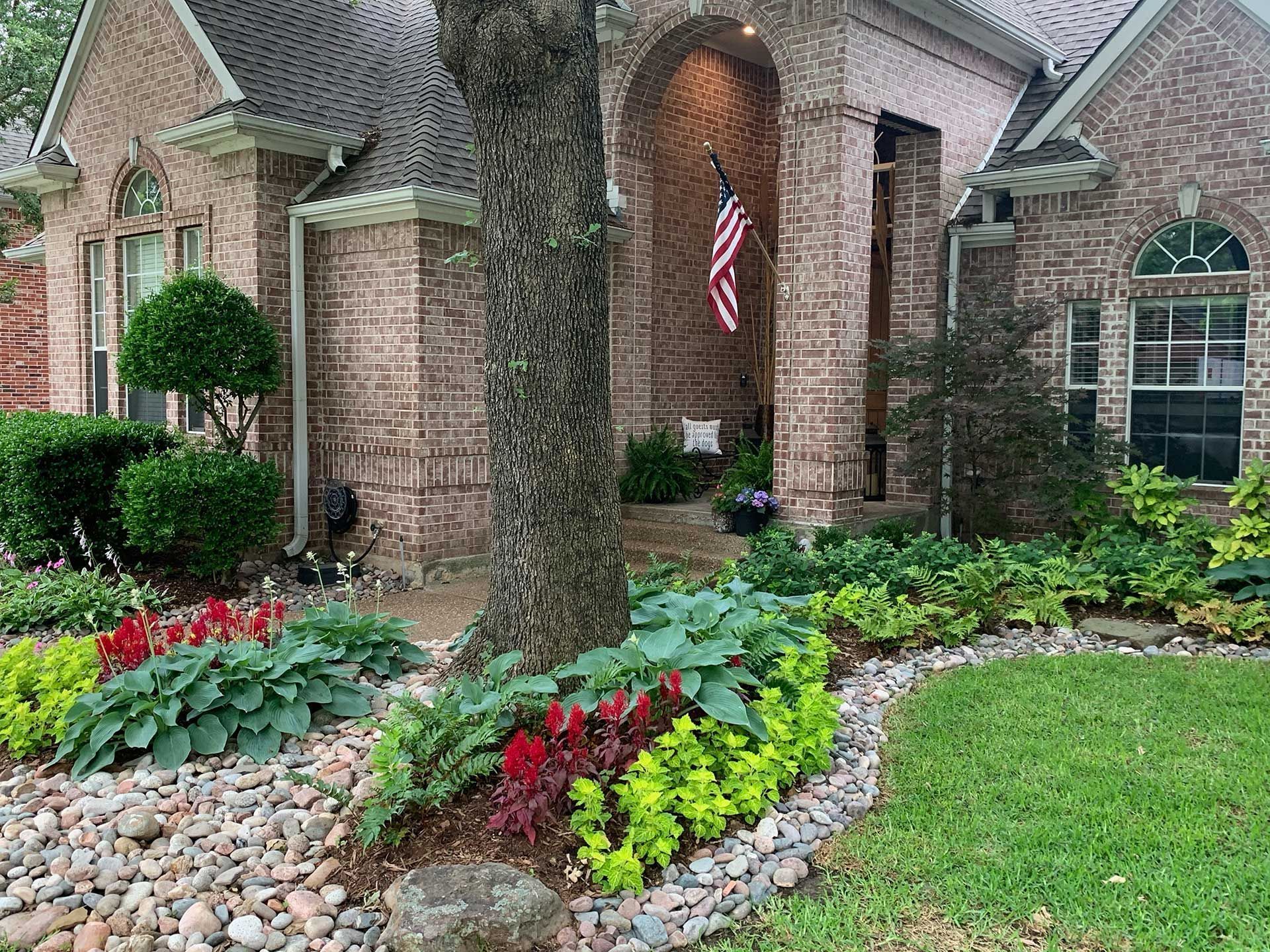 Brick house entrance with arched porch, American flag, and landscaped flower bed with colorful plants.