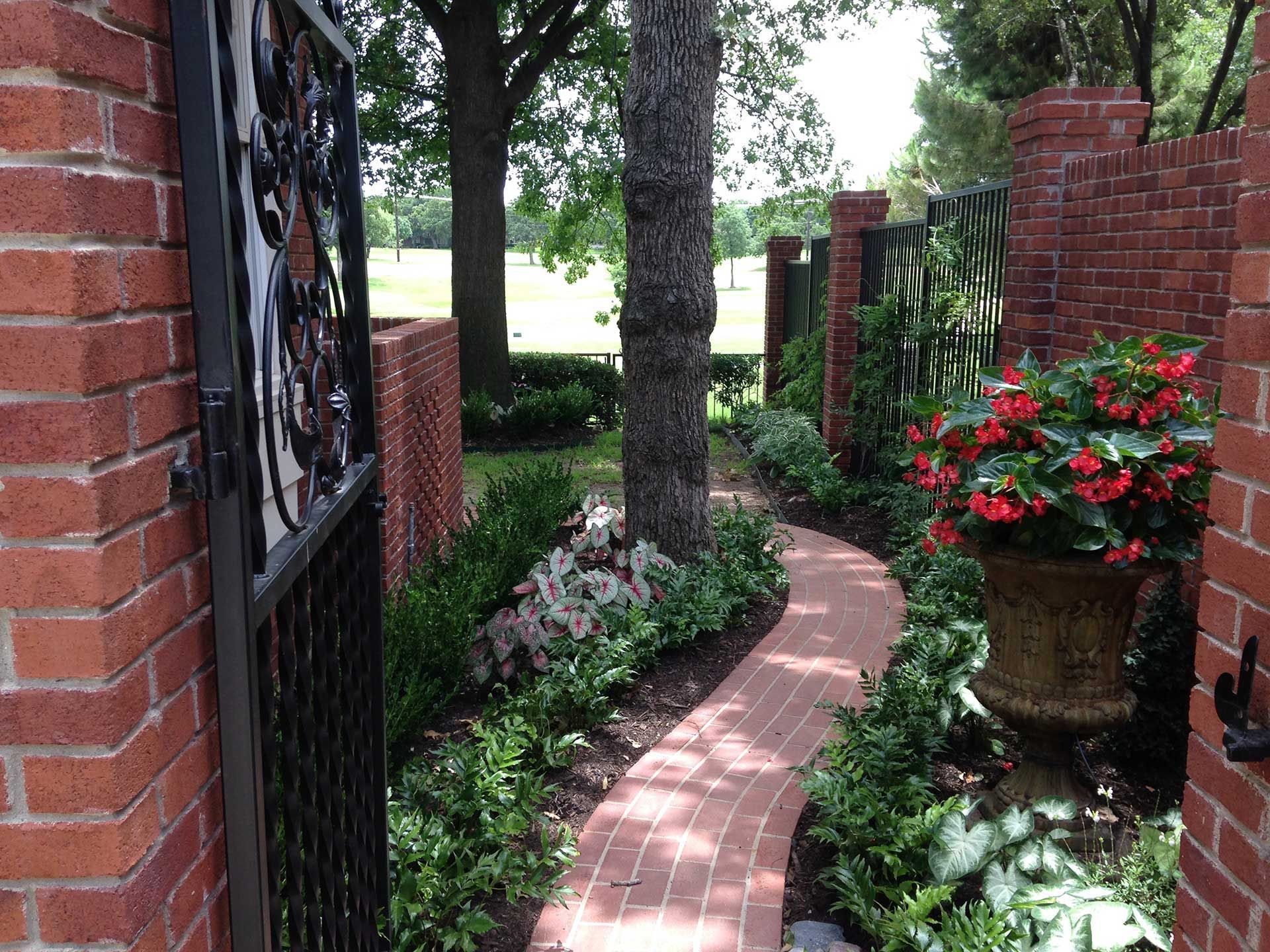 Brick garden path curving through a lush courtyard with trees, flowers, and brick walls