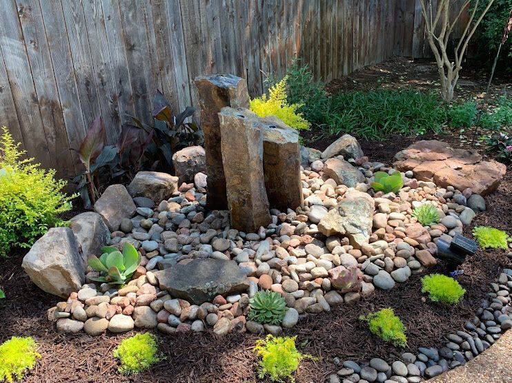 Zen garden with stacked stone sculpture, river rocks, and small green plants in a backyard.