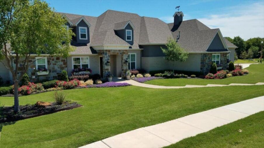 Large suburban house with a manicured green lawn, flower beds, and a curved sidewalk under a clear sky