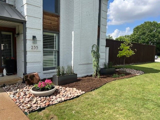 Front yard with flower bed, white house exterior, and green lawn beside a wooden fence.
