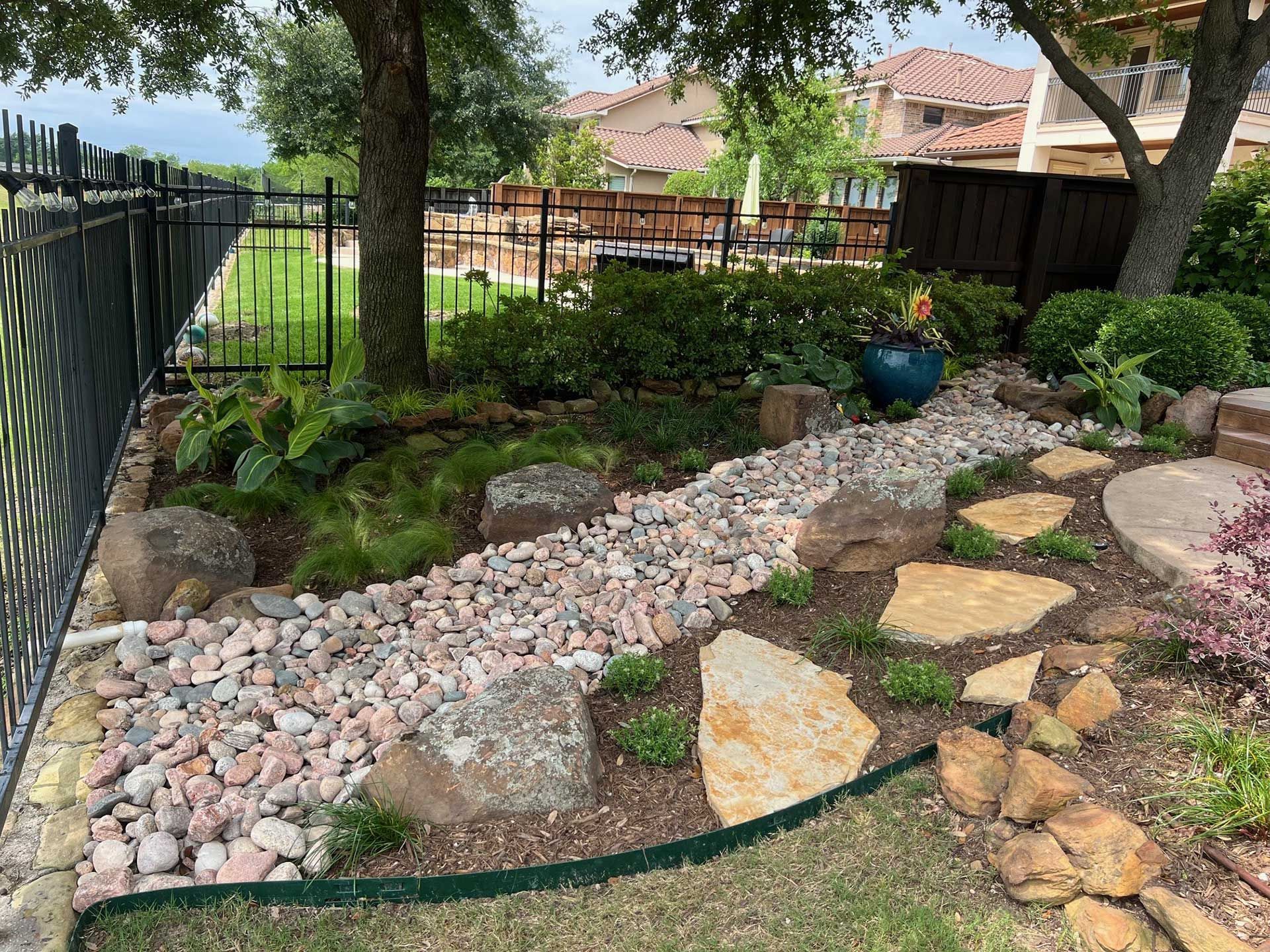 Landscaped backyard garden with rocks, winding stone path, shrubs, and a black metal fence