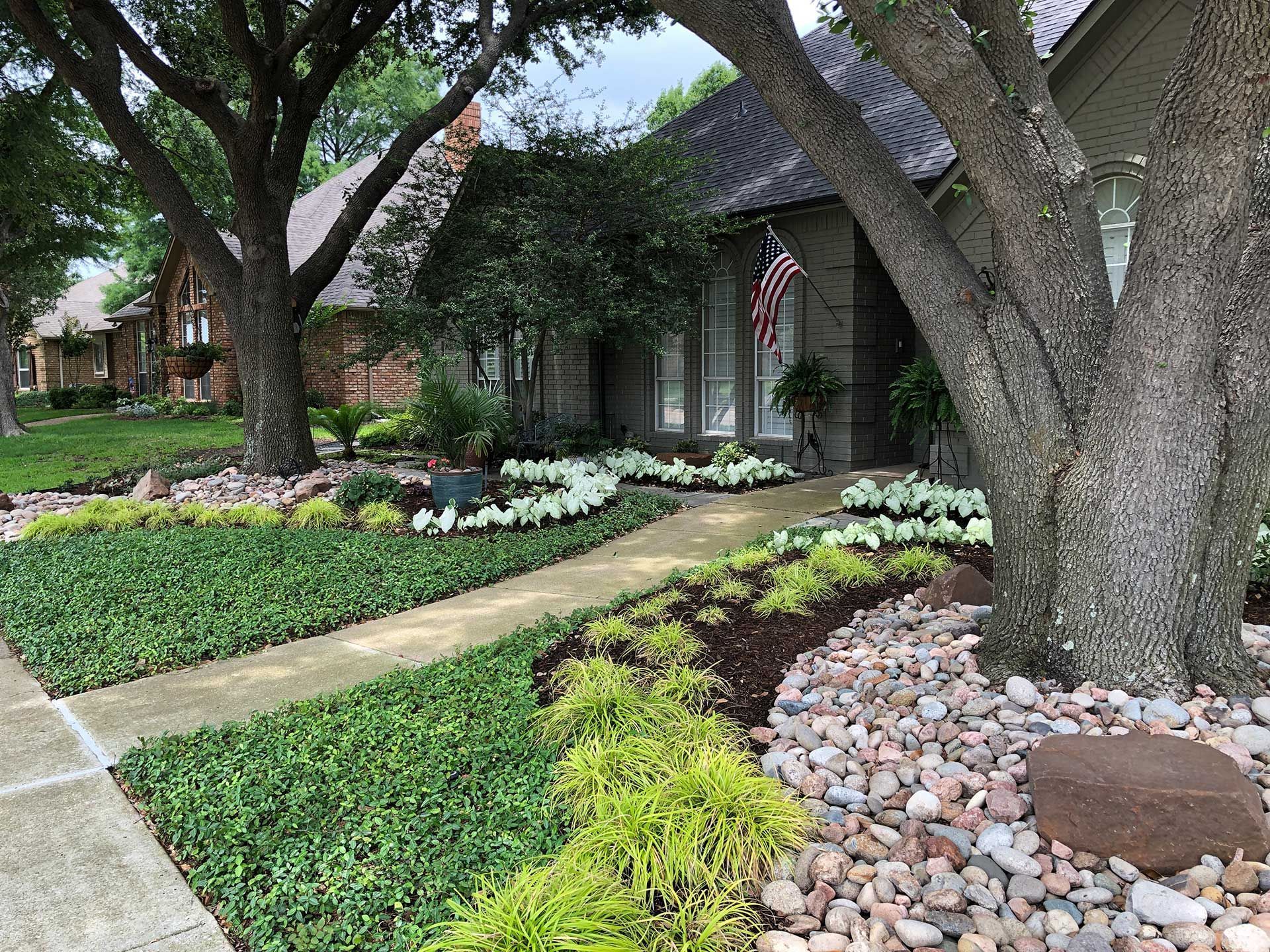 Landscaped front yard with flower beds, stone path, and large trees beside a house