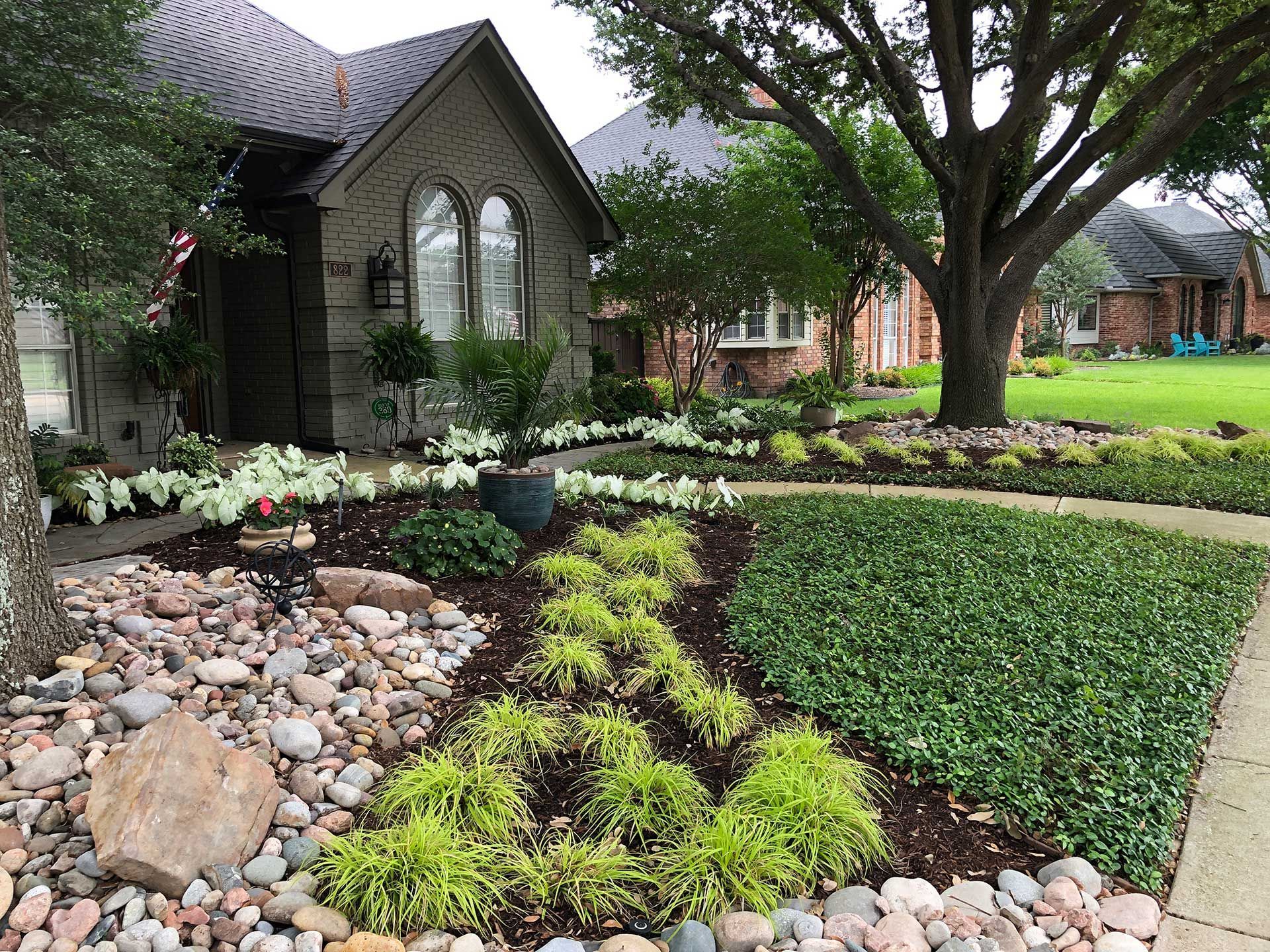 Landscaped garden with stone border, flowering beds, lawn, and a small stone building beside a large tree