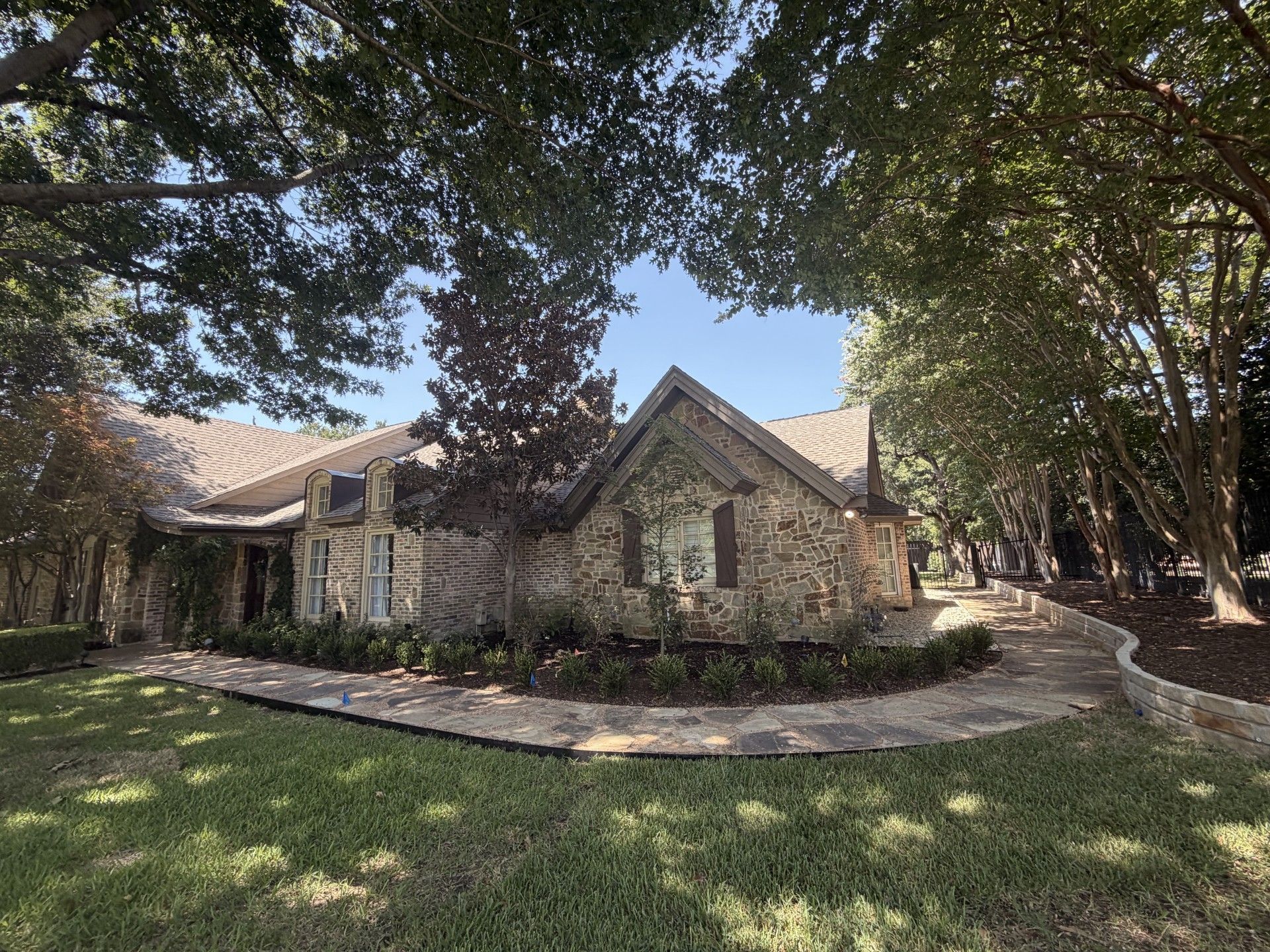Stone house with arched windows and a curved walkway under large shade trees