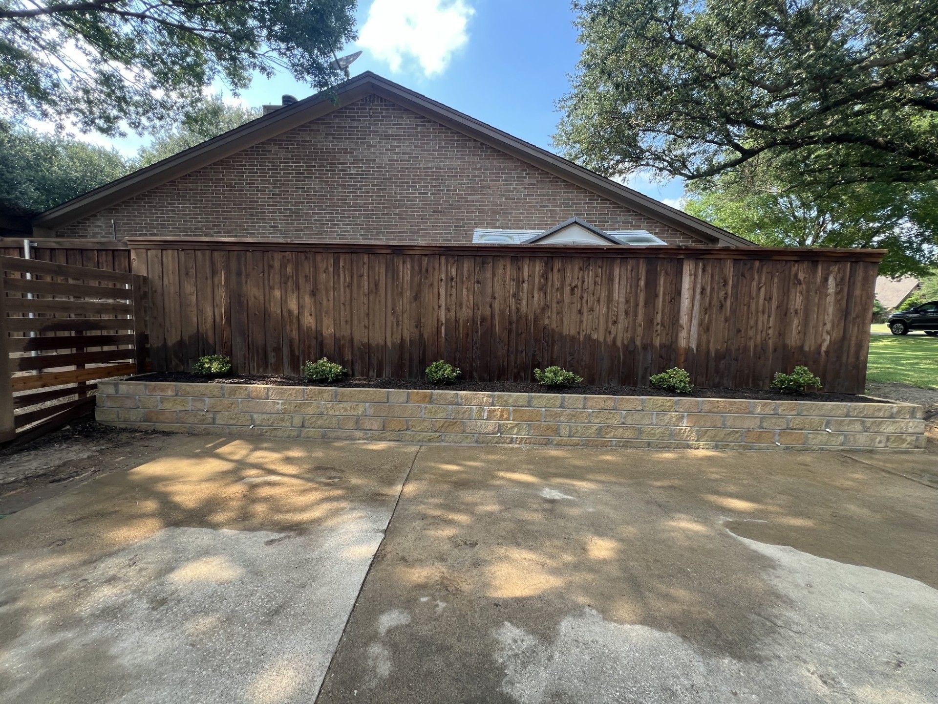 Wooden privacy fence and gate in front of a house, with trees overhead and sunlit concrete driveway