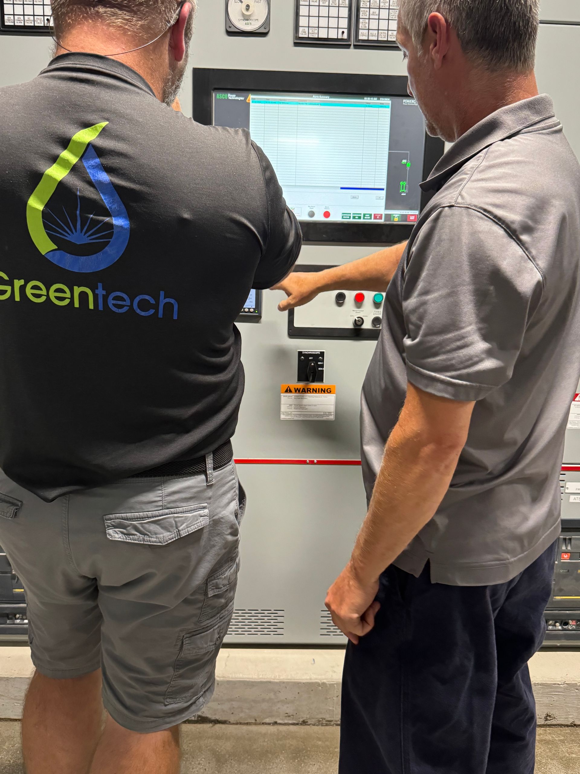 Two men are standing in front of a Greentech control panel