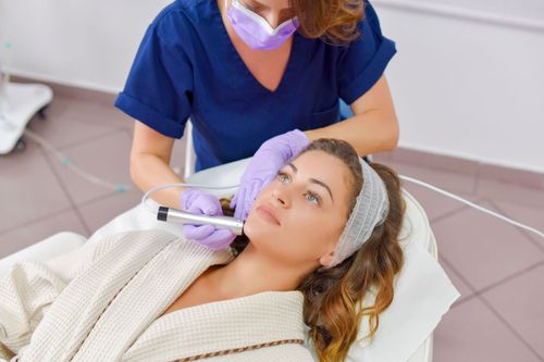 Aesthetician using a handheld device on a reclining client during a facial treatment.