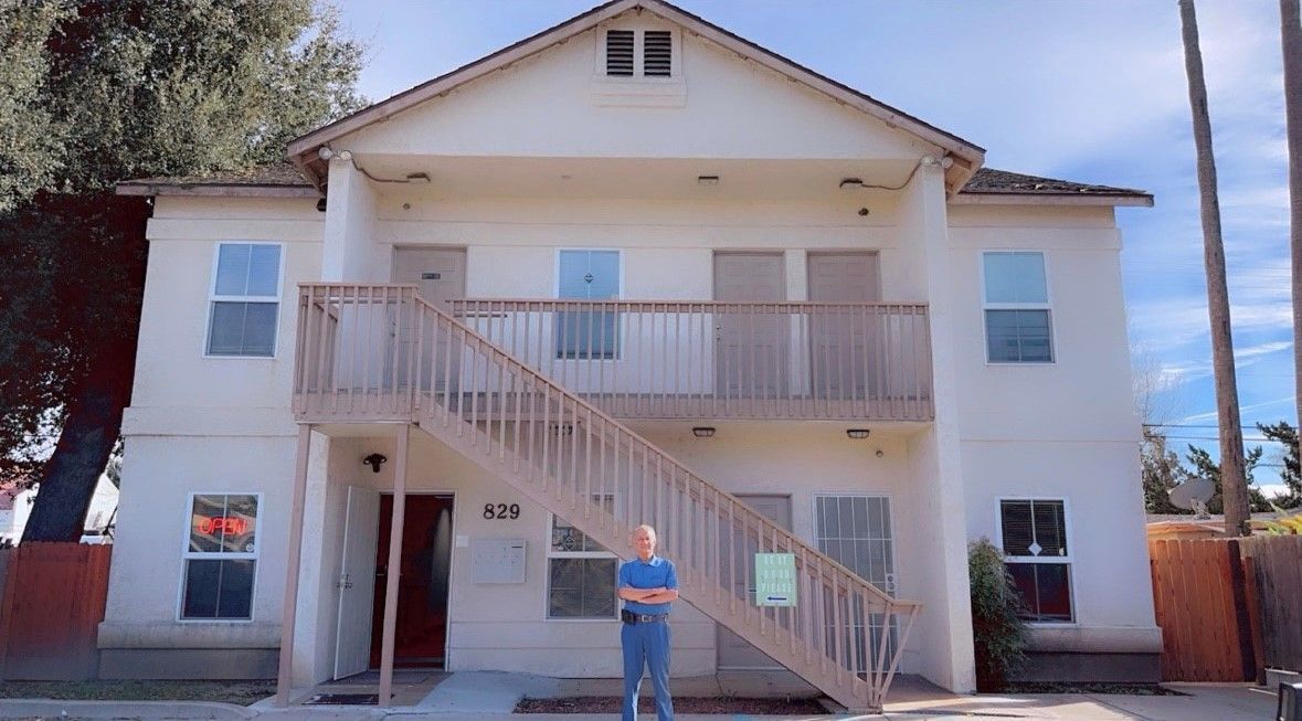 Man stands in front of a two-story white apartment building with wooden stairs, cloudy sky.