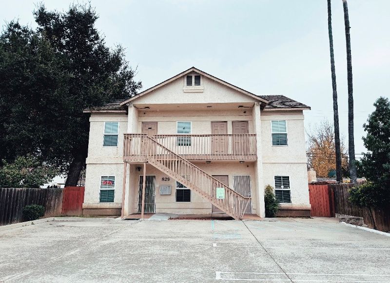 Two-story tan apartment building with wooden stairs and doors, surrounded by trees and a paved lot.