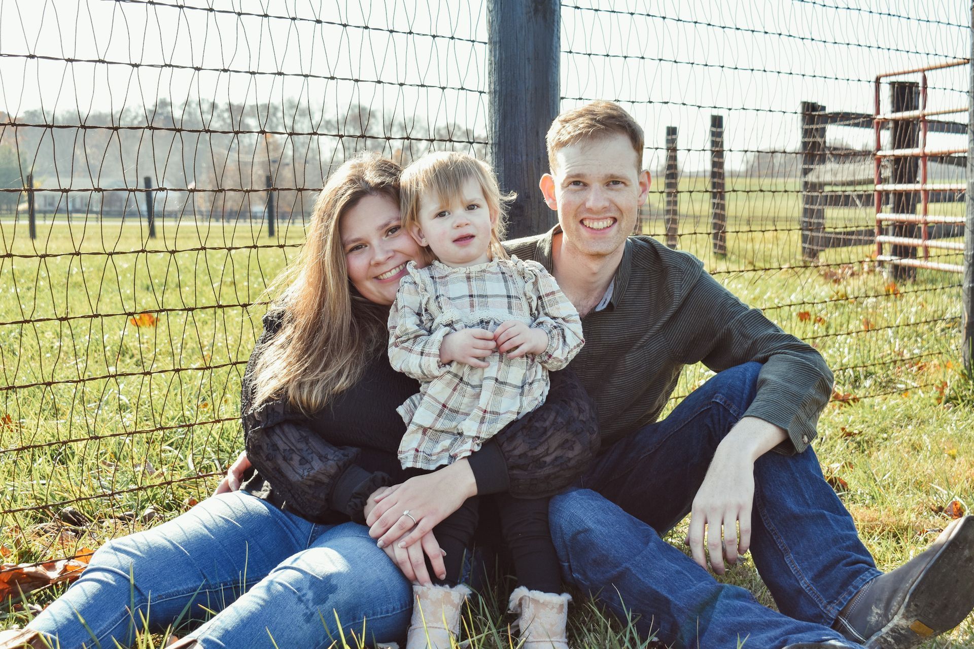 Family of three smiling, sitting on grass, in front of a wire fence and wooden gate.