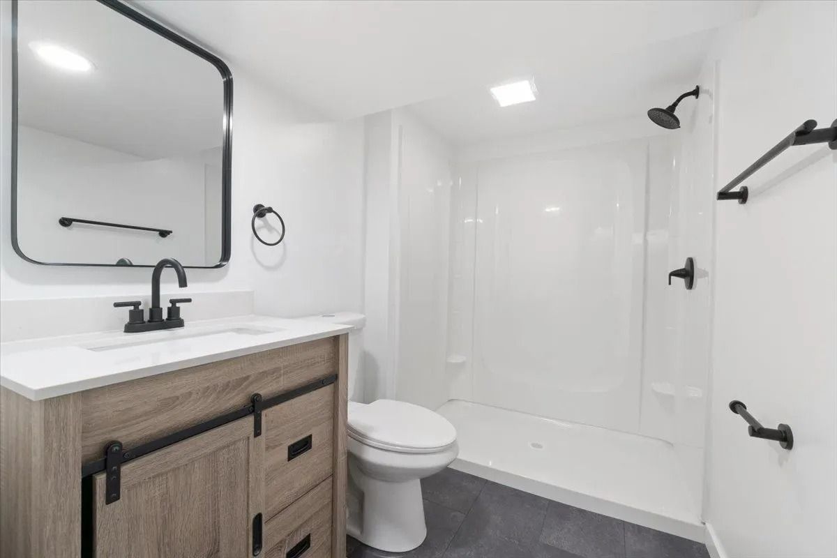 Bathroom with white walls and a light wood vanity, shower, toilet, and black fixtures.